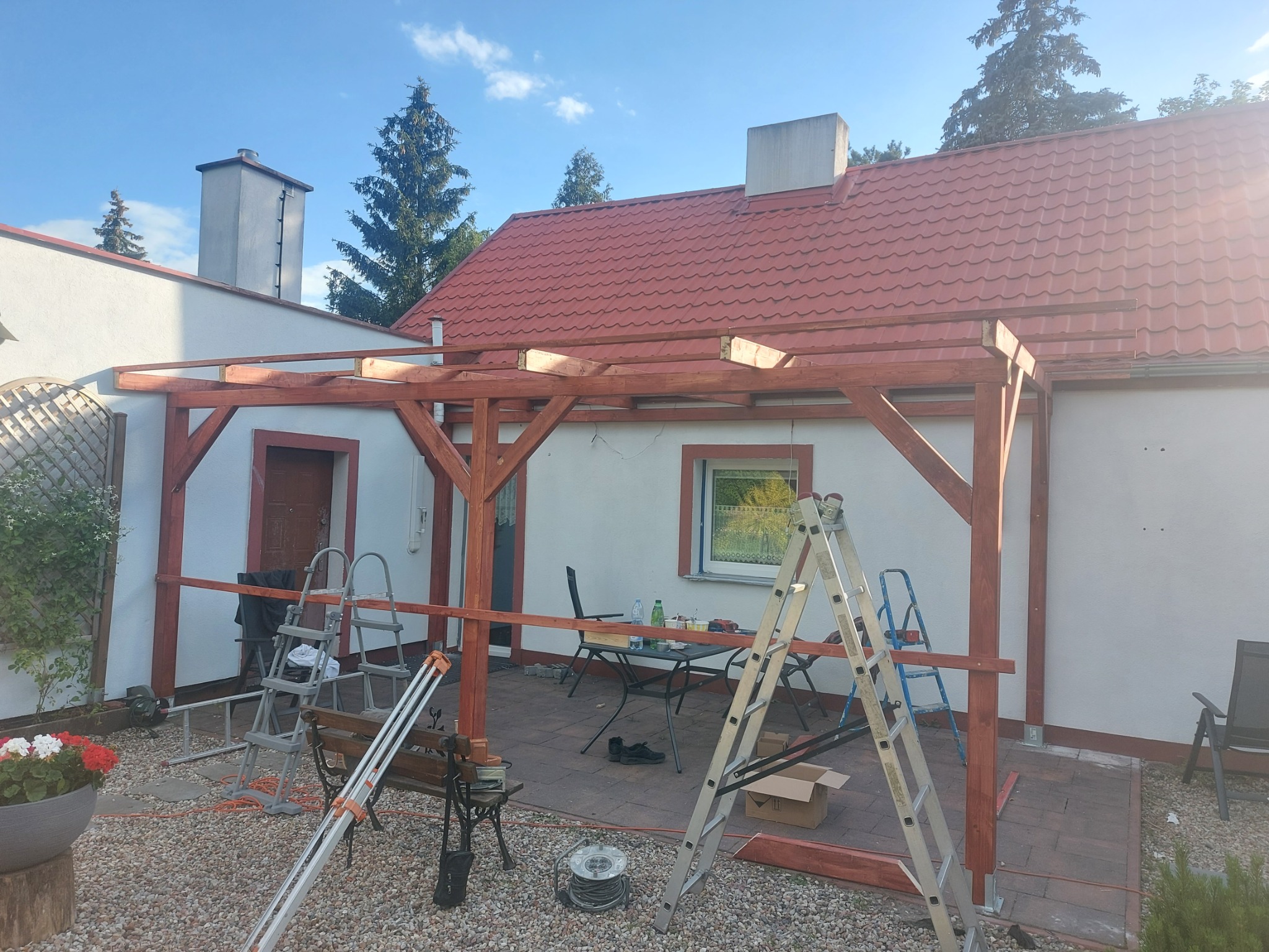 Drewniana konstrukcja zadaszenia tarasu w trakcie budowy, widoczne drabiny, narzędzia i częściowo zadaszony dom z czerwoną dachówką w tle.