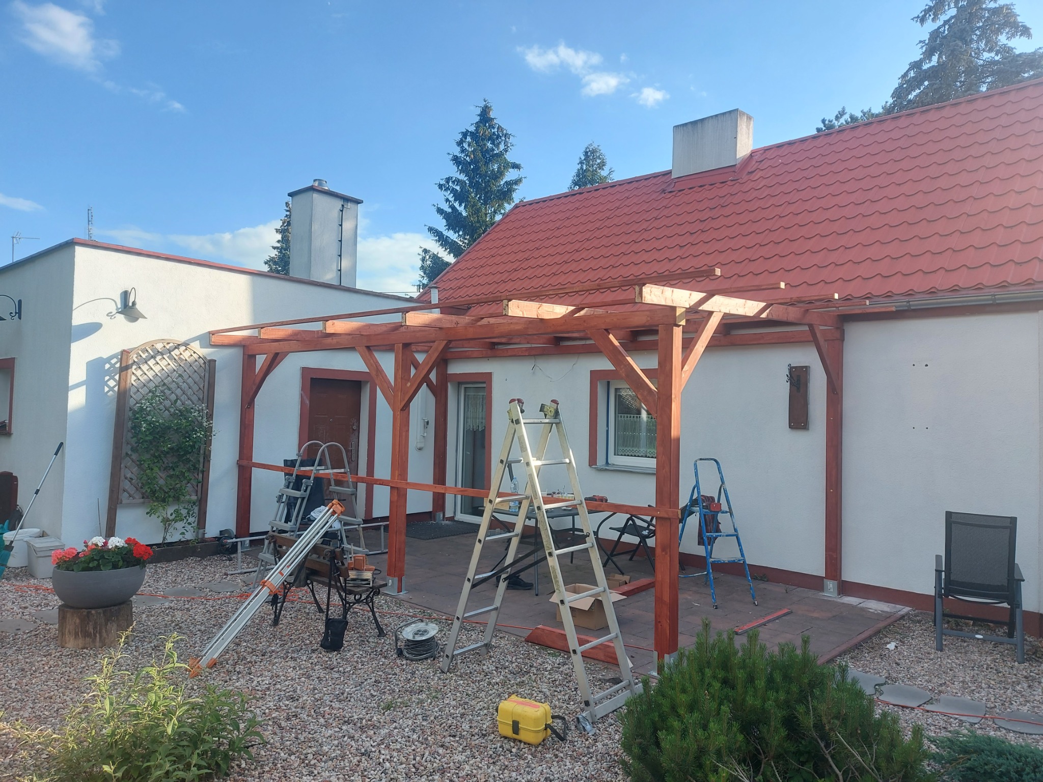 Montaż drewnianej pergoli przy domu z czerwoną dachówką. Widoczne drabiny i narzędzia. Prace w toku, słoneczny dzień.