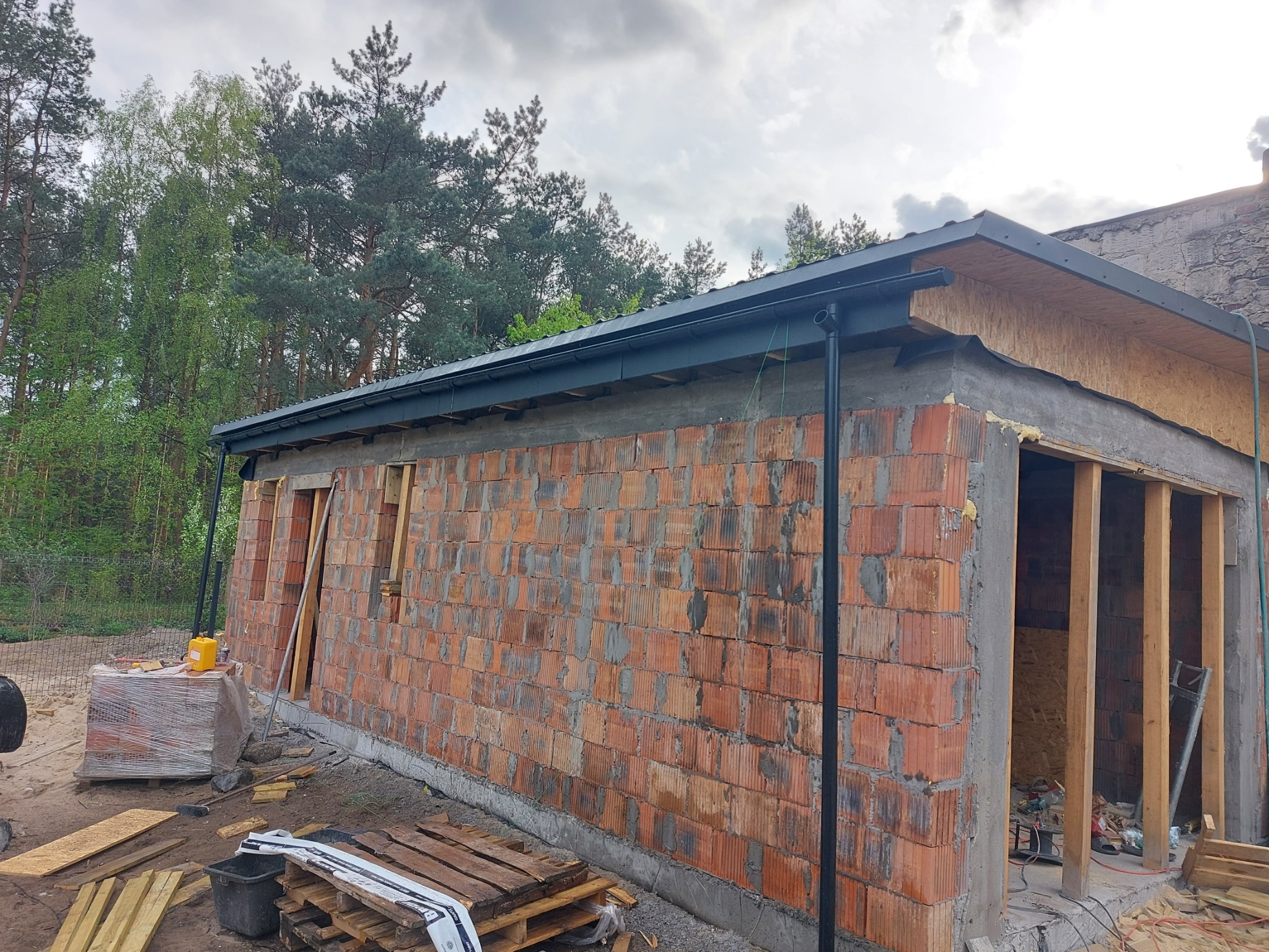 Budowa domu w trakcie: murowane ściany z pustaków ceramicznych, zamontowane rynny, widoczna więźba dachowa. Elementy konstrukcyjne i materiały budowlane na placu budowy.