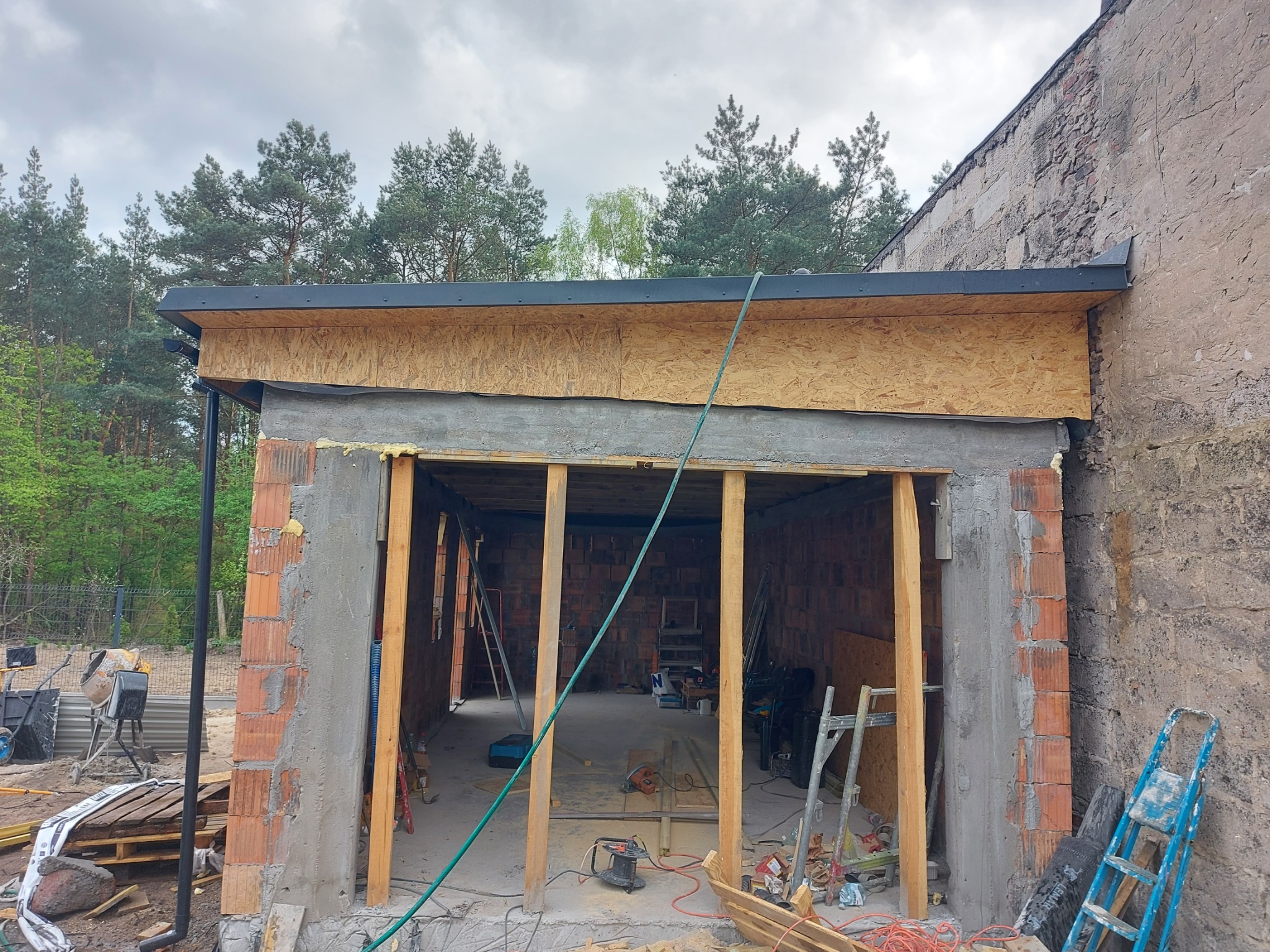 Surowy stan rozbudowy domu z widoczną konstrukcją dachu, ścianami z cegieł i elementami drewnianymi. Wnętrze z narzędziami budowlanymi i materiałami.