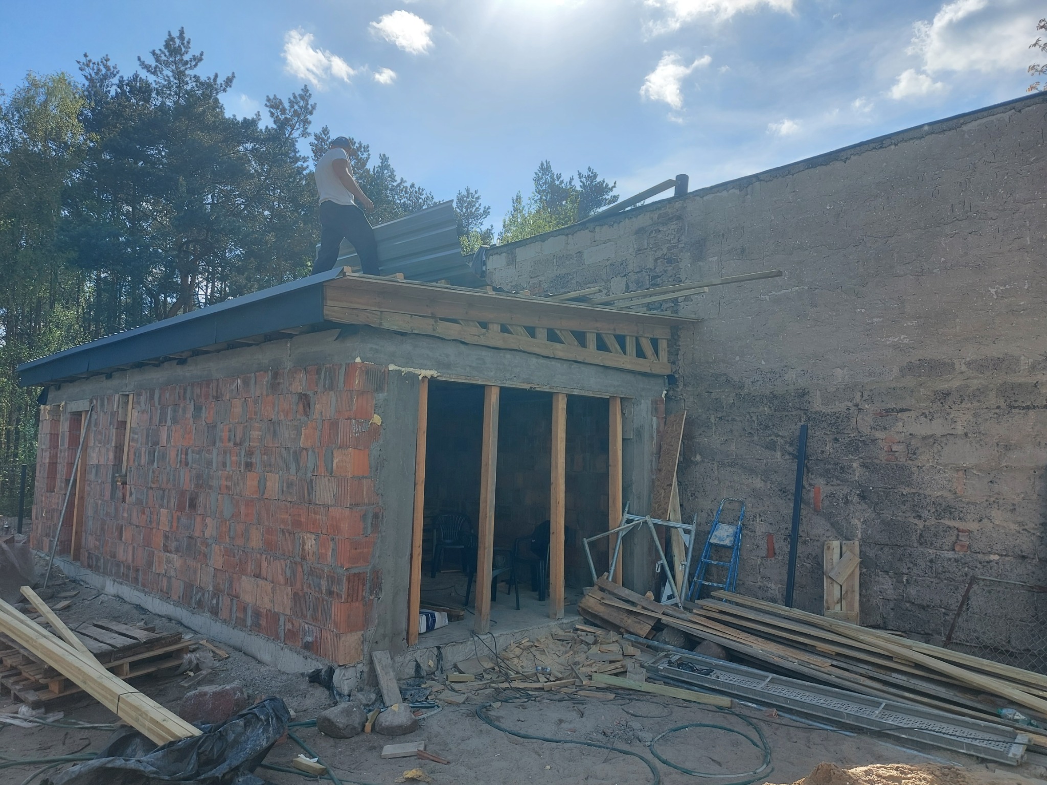 Budowa garażu z cegły: murarz układa blachę na dachu. Widoczne drewniane elementy konstrukcyjne, narzędzia i materiały budowlane w otoczeniu drzew.