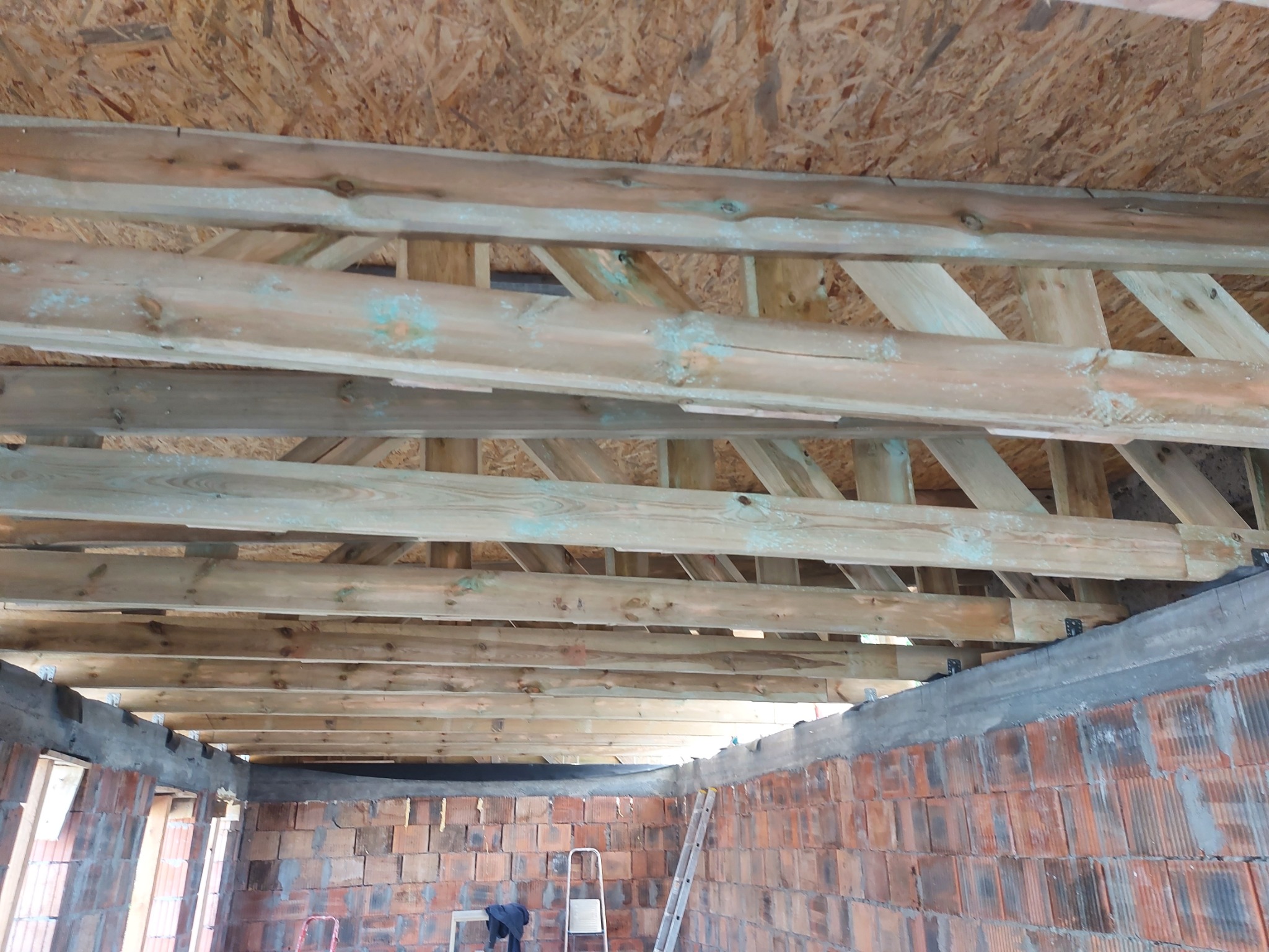 Wnętrze budynku w trakcie budowy. Widoczna drewniana więźba dachowa z impregnacją, murowane ściany z czerwonej cegły i fragment drabiny.