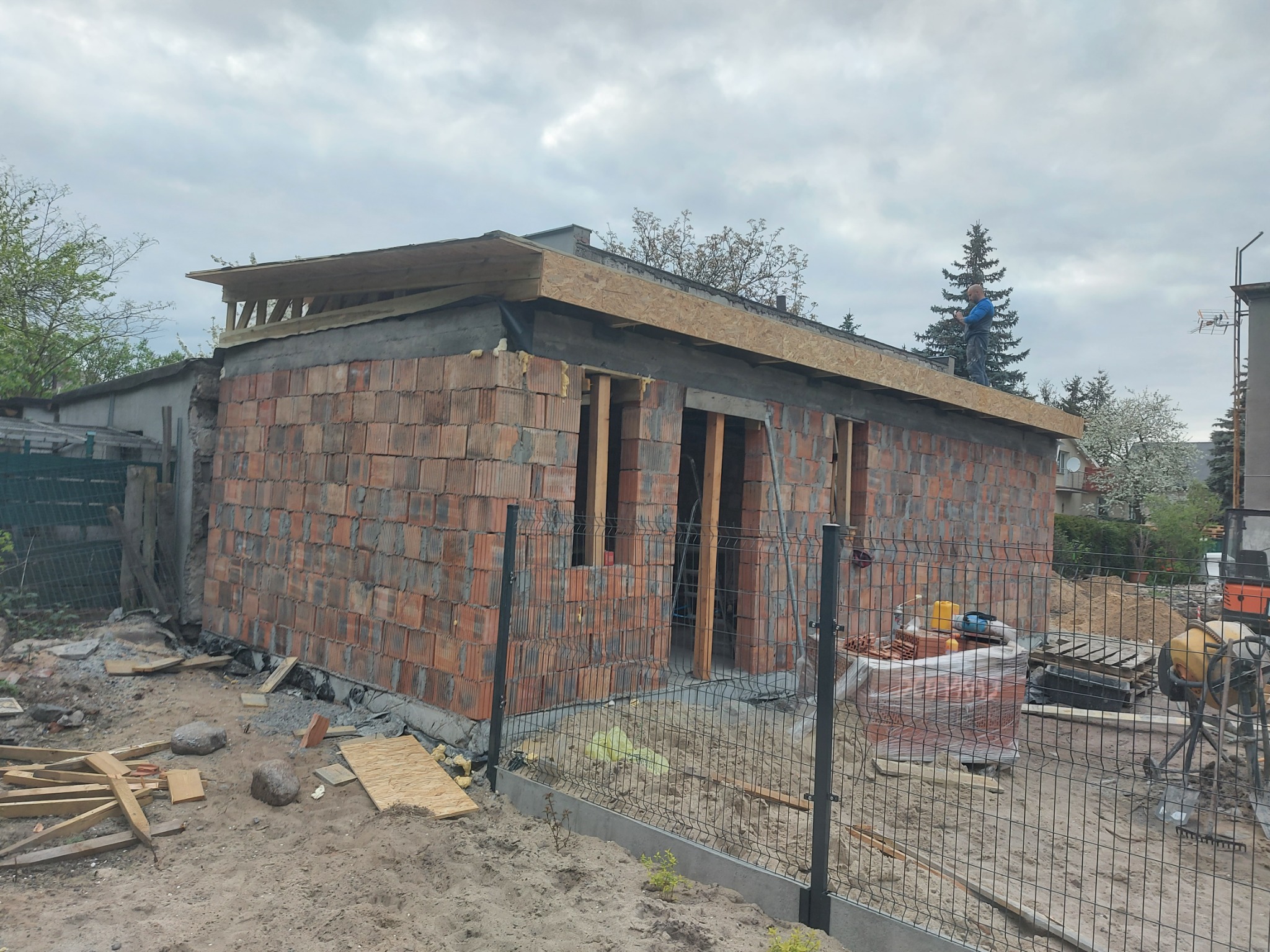 Murowany budynek w trakcie budowy z drewnianą konstrukcją dachu, widoczny pracownik na dachu, teren budowy z materiałami i ogrodzeniem panelowym.