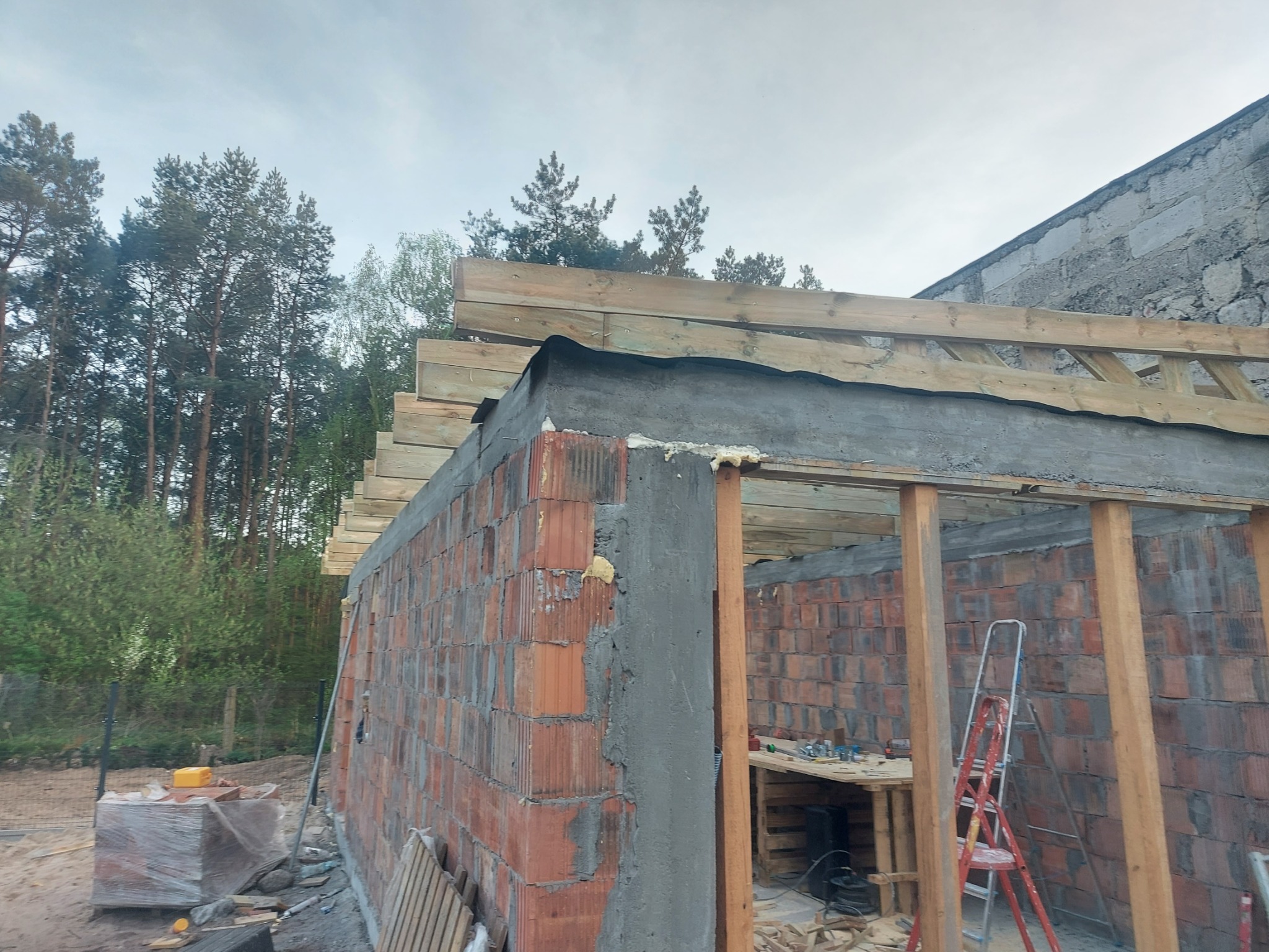 Budowa niskiego budynku z czerwonej cegły, widoczne drewniane elementy konstrukcyjne dachu i narzędzia na stole wewnątrz. W tle las i fragment budynku z szarego kamienia.