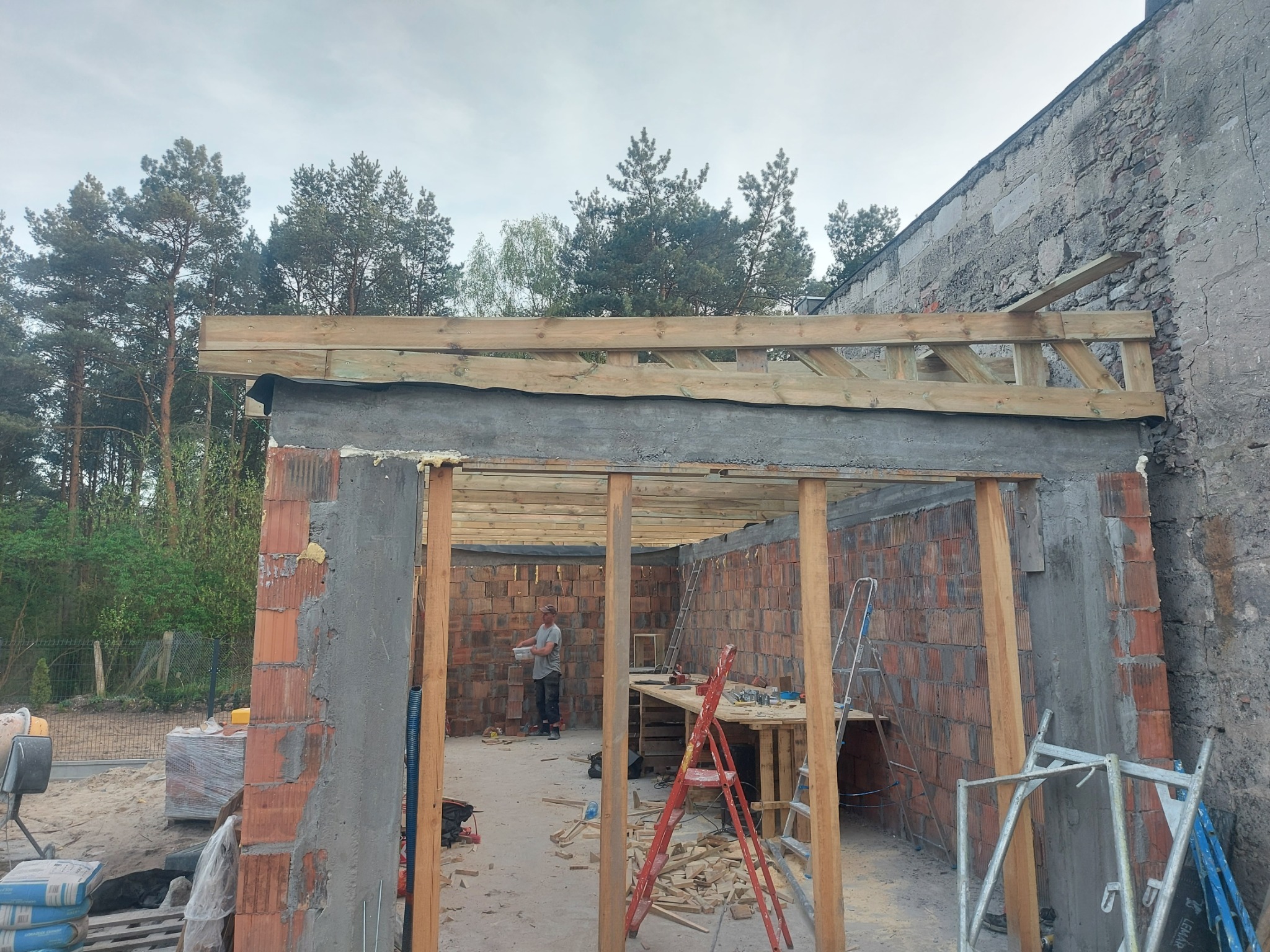 Budowa garażu z czerwonej cegły: wzniesione mury, drewniana konstrukcja dachu w trakcie montażu, drabiny i narzędzia budowlane wewnątrz. Praca w toku.