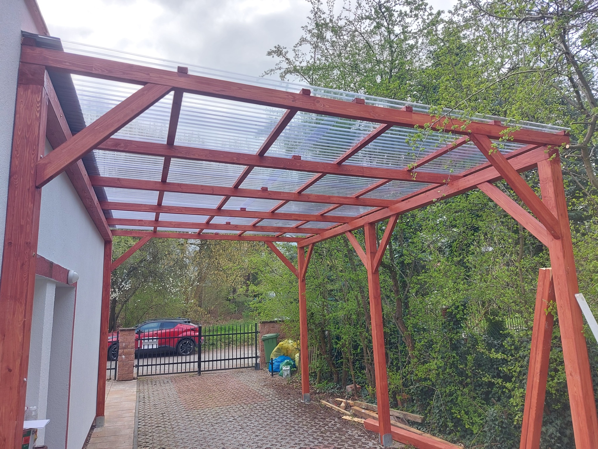 Drewniana wiata z przezroczystym zadaszeniem, widok na podjazd z kostki, ogrodzenie i zieleń. Konstrukcja pomalowana na czerwono-brązowy kolor.