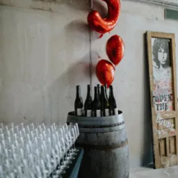 Dekoracja na przyjęcie: balony w kształcie cyfry 5, wino w drewnianej beczce, rząd kieliszków. W tle betonowa ściana i drzwi z graffiti.