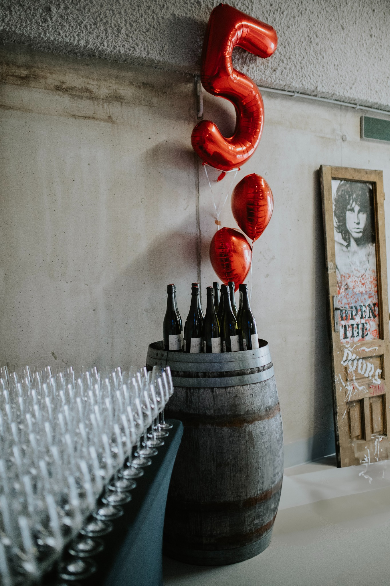 Dekoracja na przyjęcie: balony w kształcie cyfry 5, wino w drewnianej beczce, rząd kieliszków. W tle betonowa ściana i drzwi z graffiti.