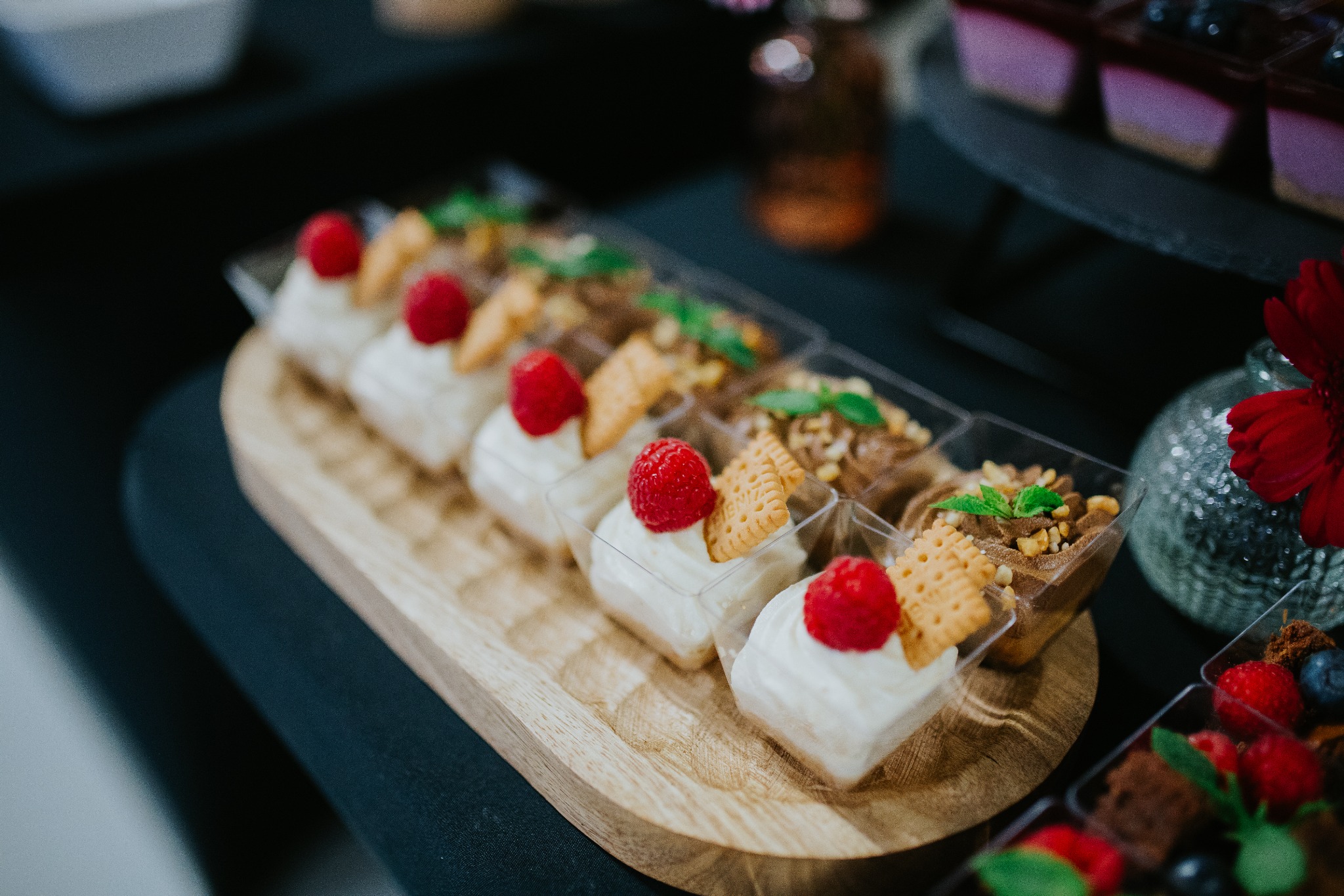 Deserowe mini porcje: kremowe z maliną i ciasteczkiem, czekoladowe z orzechami, owocowe z brownie, ułożone na drewnianej tacy. Elegancki catering.