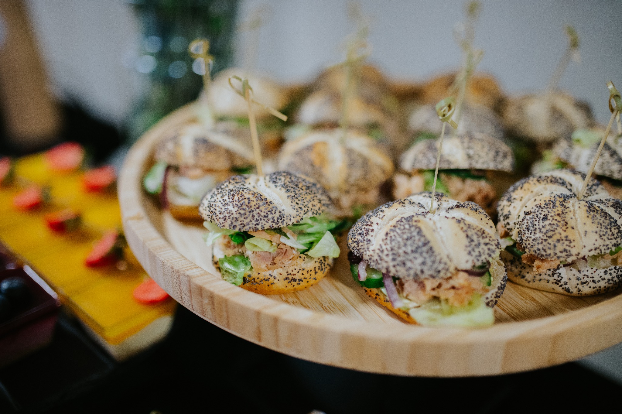 Drewniana taca z mini burgerami z ziarnistymi bułkami, sałatą i łososiem. Przekąski na przyjęcie, widoczne truskawki na żółtej desce.