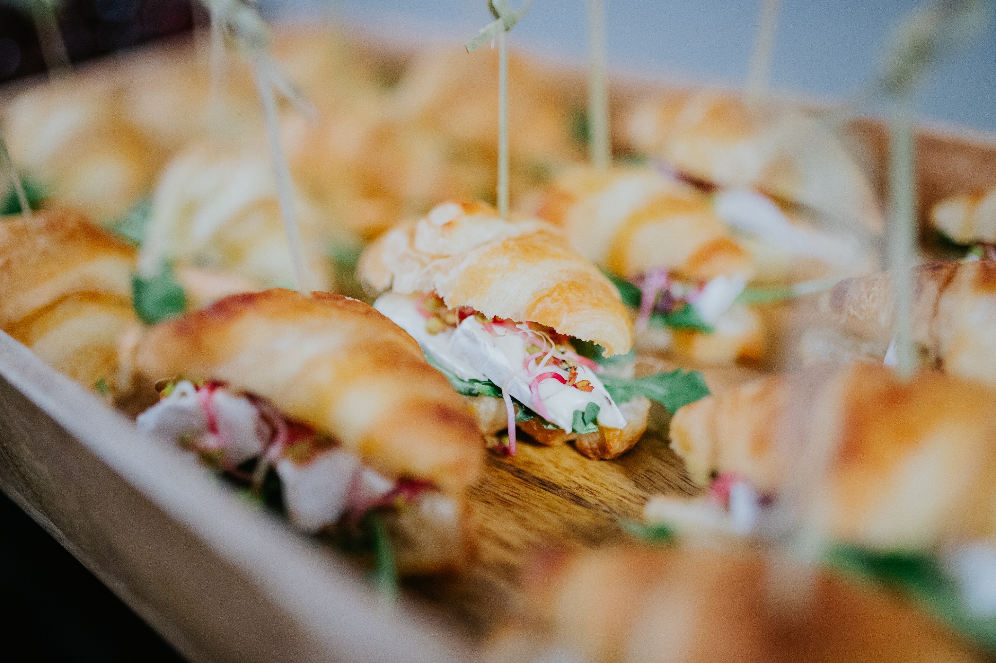 Kruche rogaliki z wykwintnym nadzieniem na drewnianej desce, ozdobione zielonymi listkami i spięte ozdobnymi wykałaczkami. Idealne na elegancki catering.