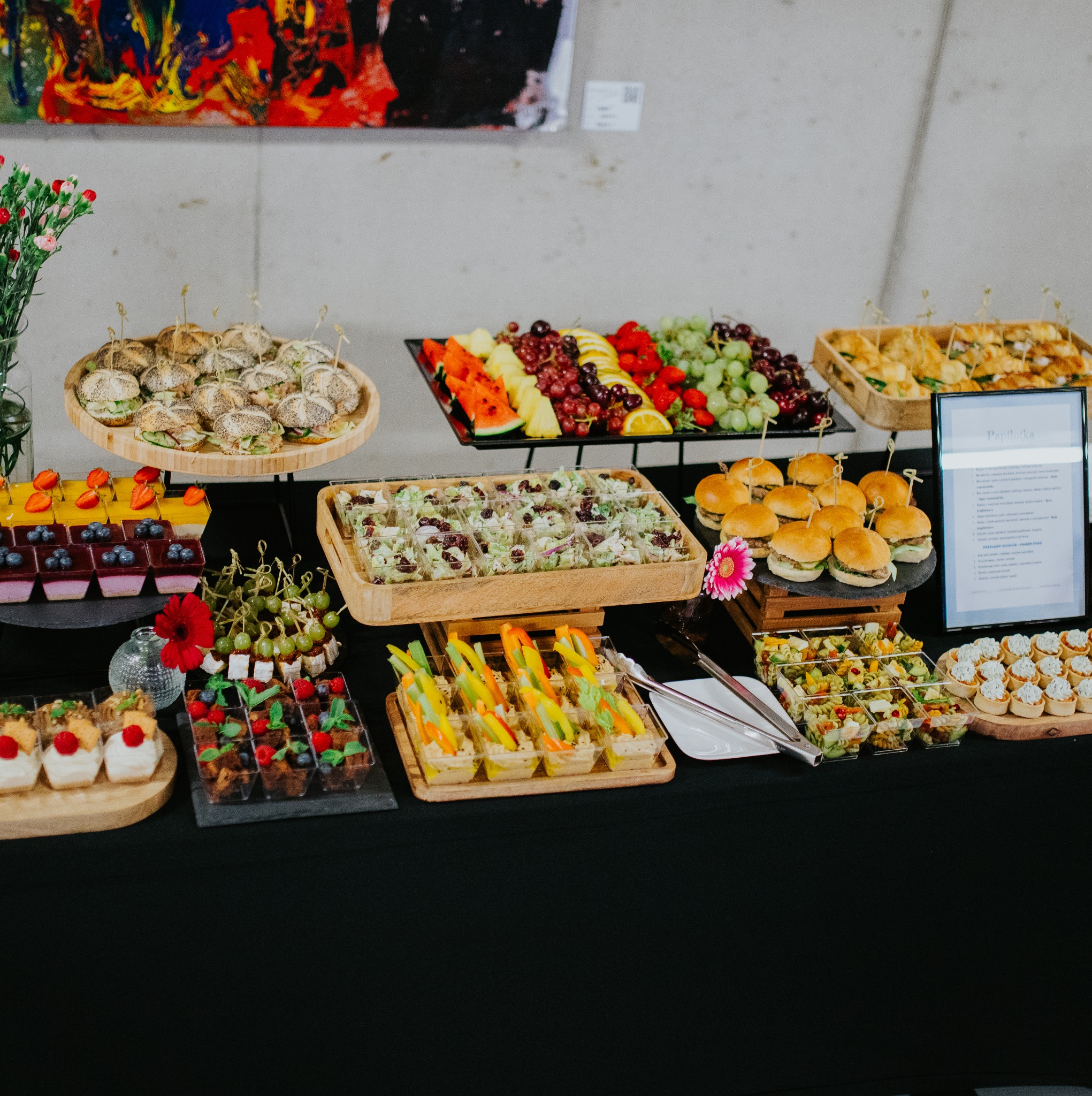 Stół cateringowy z różnorodnymi przekąskami: mini burgery, sałatki w pojemnikach, owoce, desery. Elegancka prezentacja na czarnym obrusie, idealna na event firmowy.
