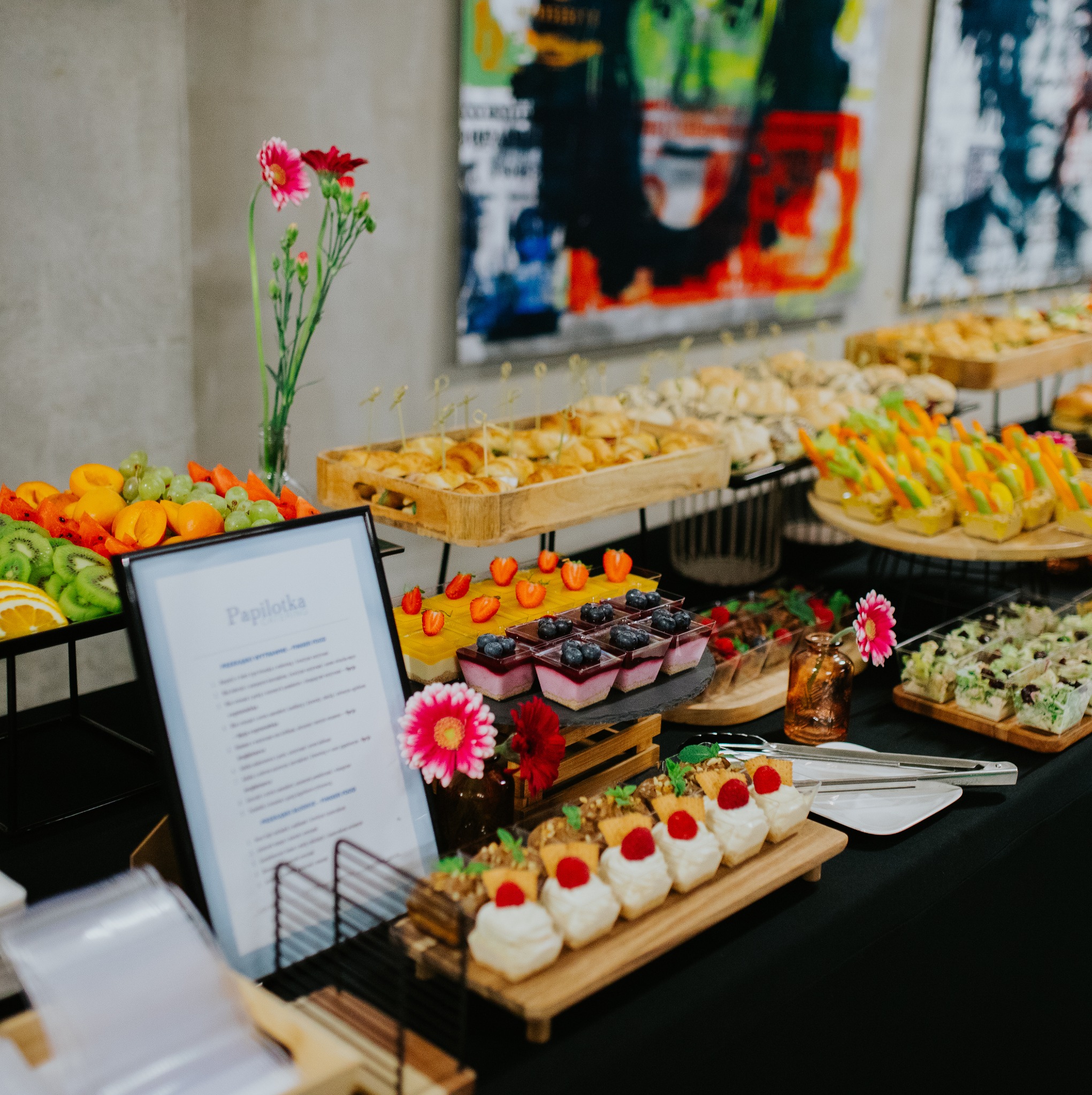 Stół cateringowy z różnorodnymi przekąskami: owoce, desery, mini kanapki. W tle abstrakcyjne obrazy i kwiaty w wazonie. Elegancka prezentacja.