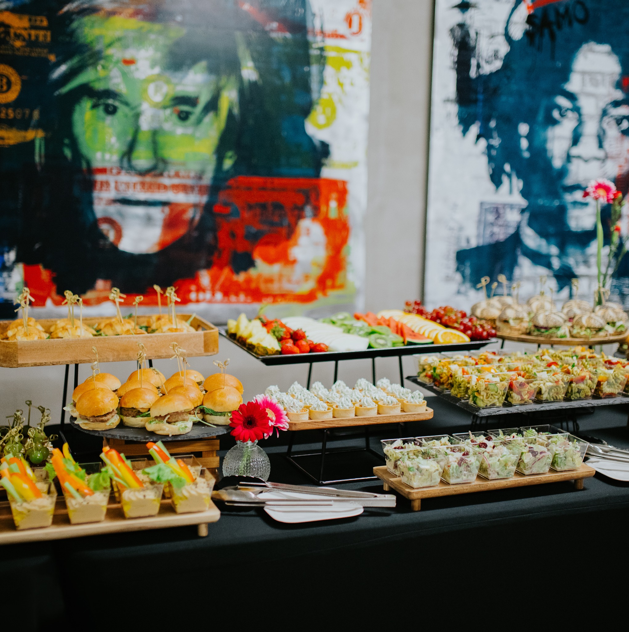 Bogato zastawiony stół cateringowy z mini burgerami, sałatkami i owocami, z abstrakcyjnymi obrazami w tle. Elegancki bufet na imprezę.