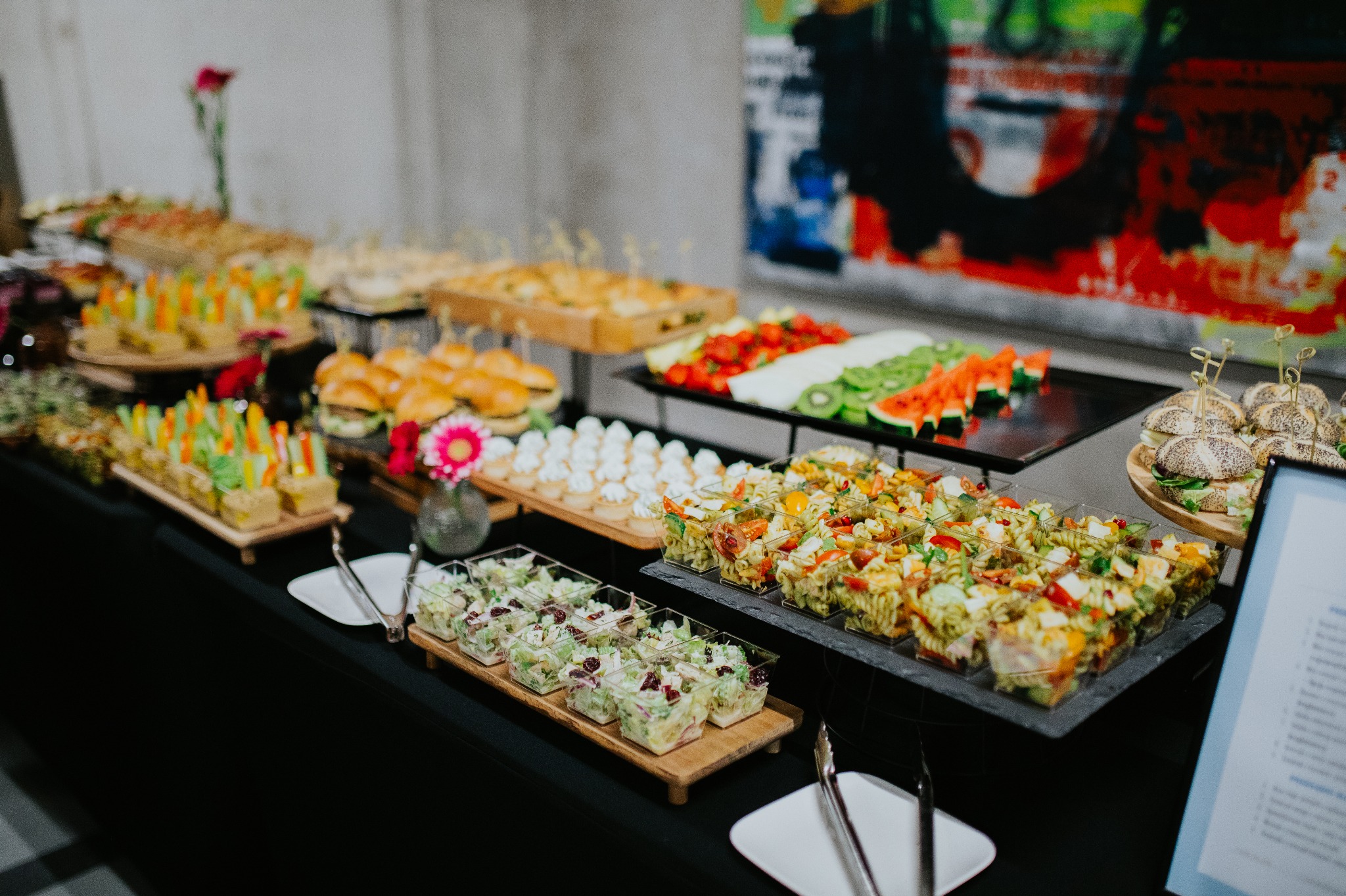 Bogato zastawiony stół cateringowy z różnorodnymi przekąskami: mini-sałatki, koreczki warzywne, burgery i owoce. W tle abstrakcyjny obraz.