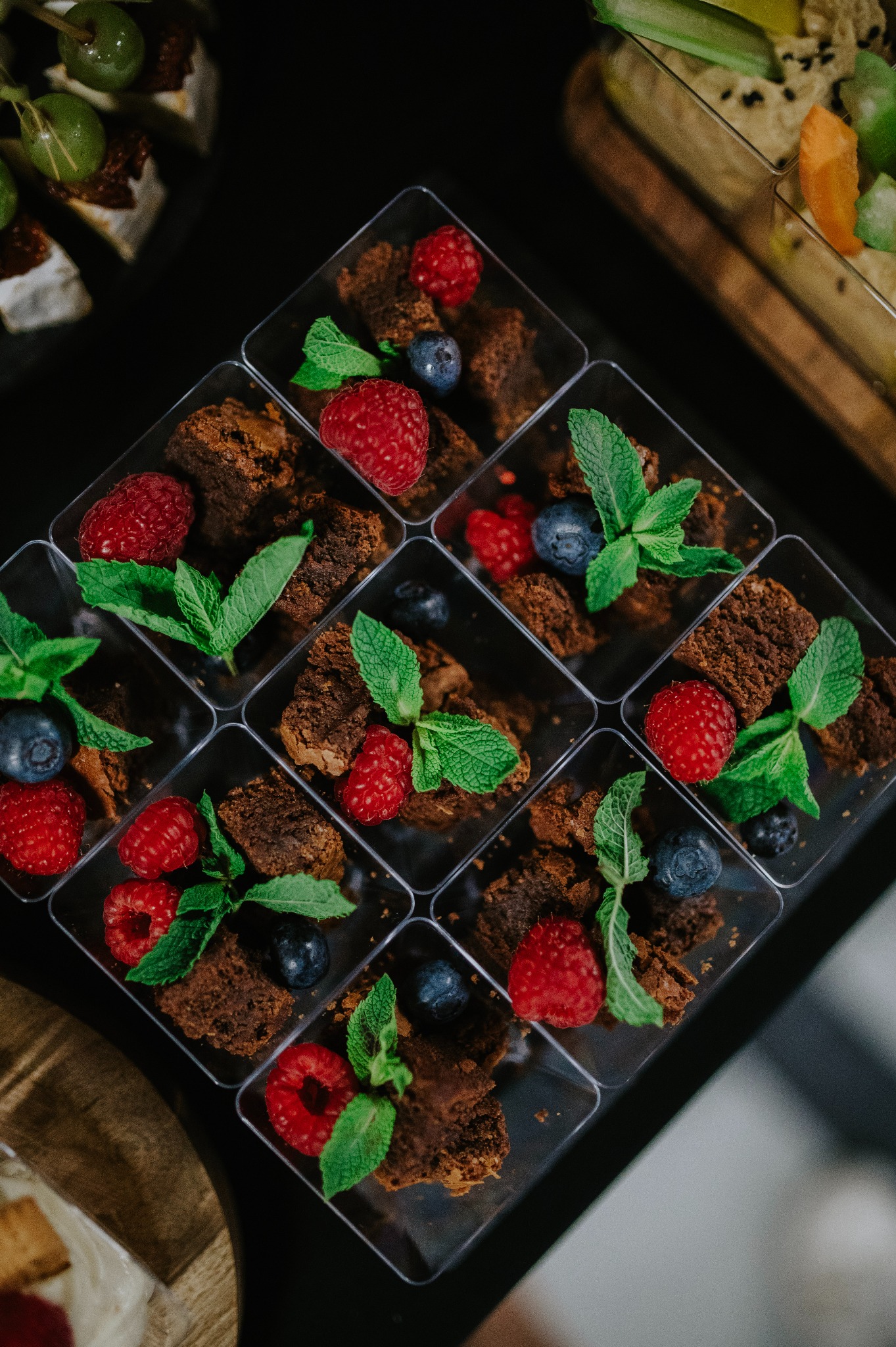 Ekskluzywne, porcjowane brownie w przezroczystych pojemnikach, udekorowane malinami, borówkami i świeżą miętą, na ciemnym tle. Elegancki catering deserowy.