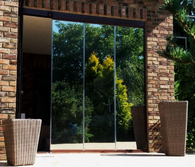 Przesuwne szklane drzwi balkonowe z przyciemnionymi szybami, odbijające zieleń ogrodu, w obramowaniu z cegły. Dwa rattanowe kosze po bokach.