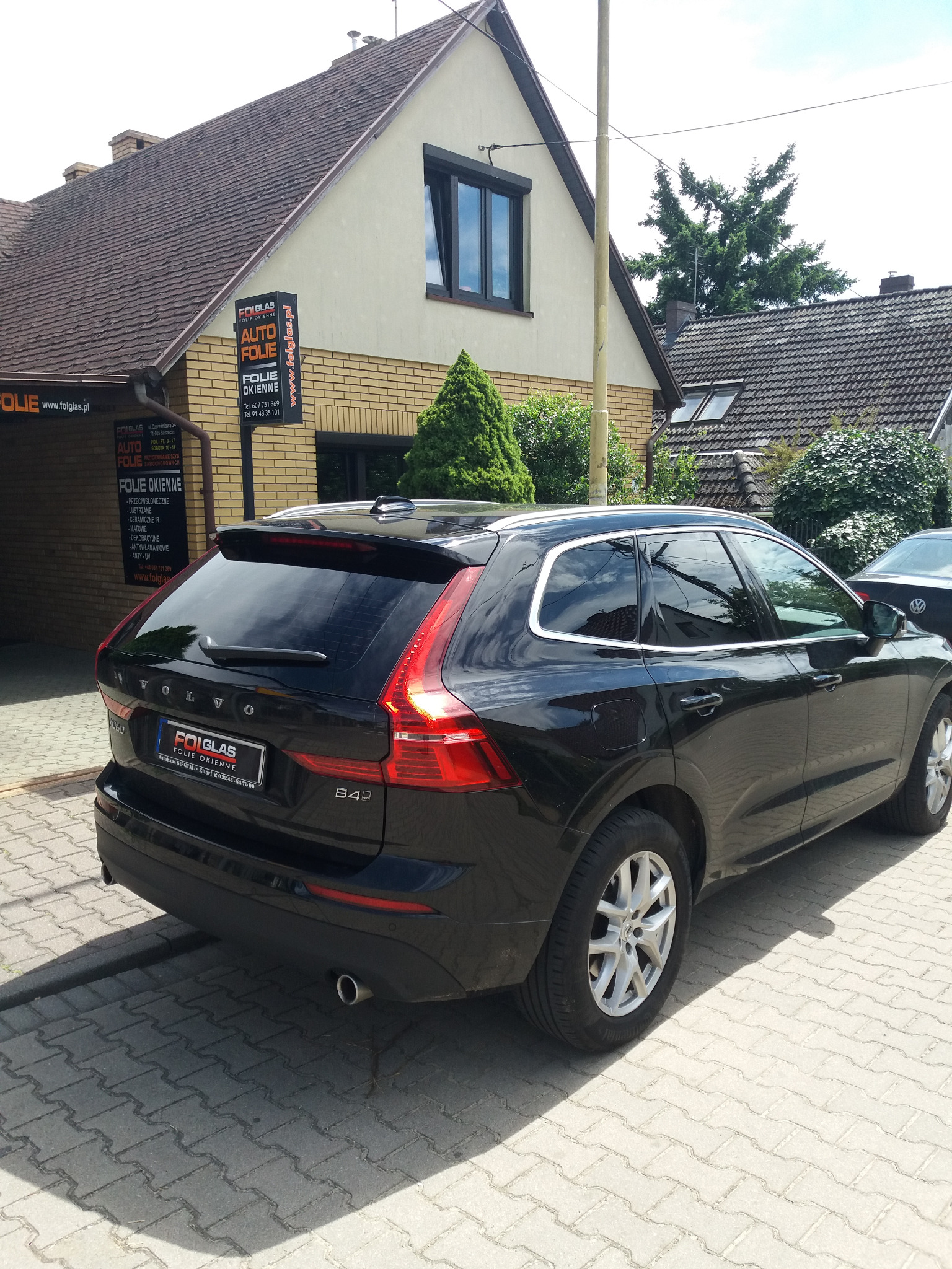 Czarne Volvo XC60 z przyciemnionymi tylnymi szybami, zaparkowane na brukowanej ulicy przed budynkiem z szyldem firmy oferującej auto folie. Widok z tyłu.