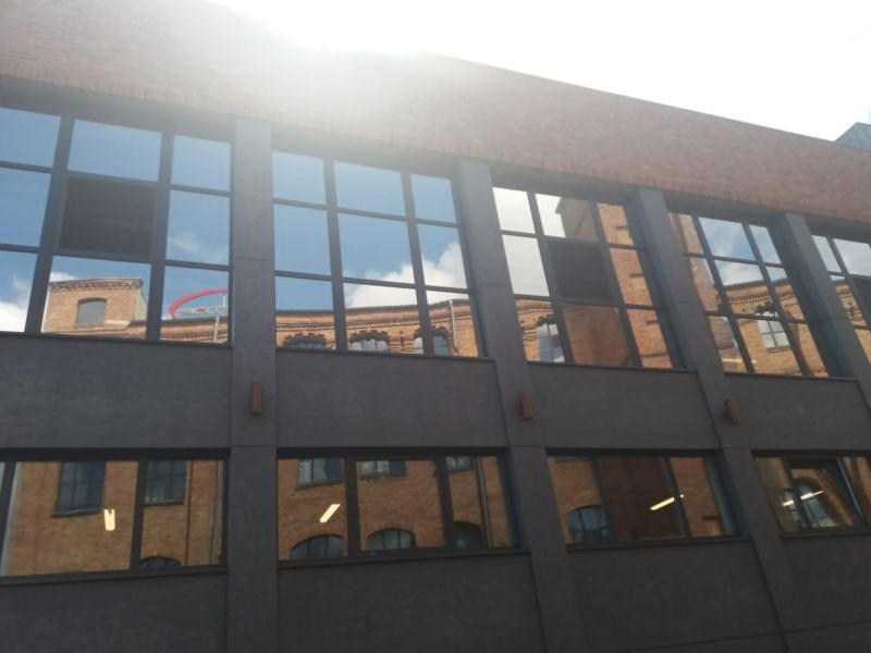 Szklana fasada budynku z efektem lustrzanego odbicia sąsiedniego budynku. Okna z ciemną folią odbijają niebo.