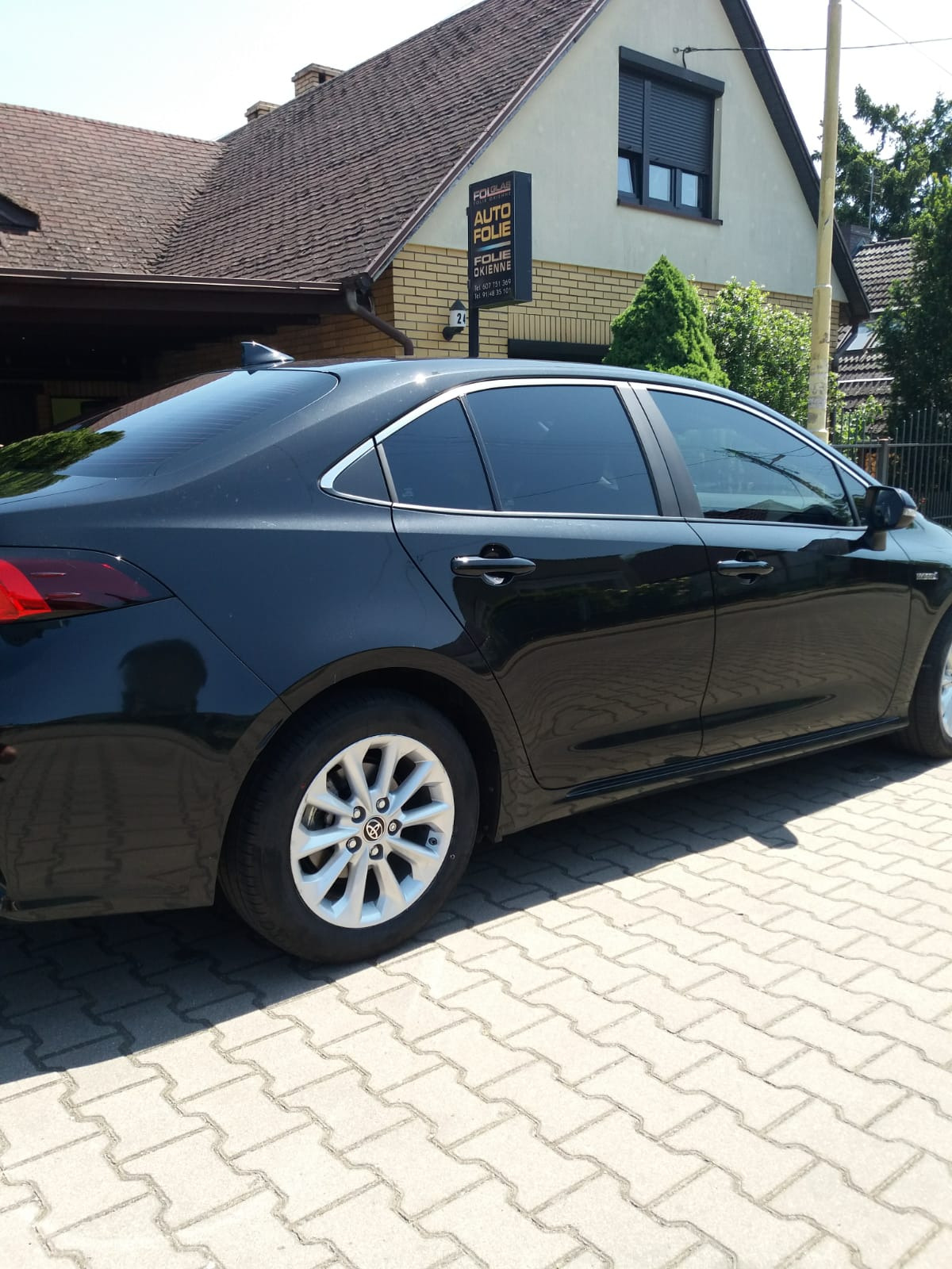 Czarny sedan z przyciemnionymi szybami, zaparkowany na kostce brukowej przed budynkiem z szyldem reklamującym usługi auto folii.