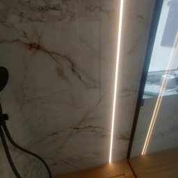 Usługi glazurnicze Wojciech Tłuchowski - Wykończona łazienka: marmurowe płytki ścienne z oświetleniem LED, odpływ liniowy w drewnopodobnej posadzce i widoczny fragment słuchawki prysznicowej.