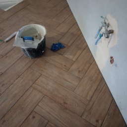 Usługi glazurnicze Wojciech Tłuchowski - Ułożona podłoga z płytek imitujących drewno w jodełkę, wiadro z zaprawą i narzędzia. W tle fragment ściany z instalacją wodną. Remont łazienki w trakcie.