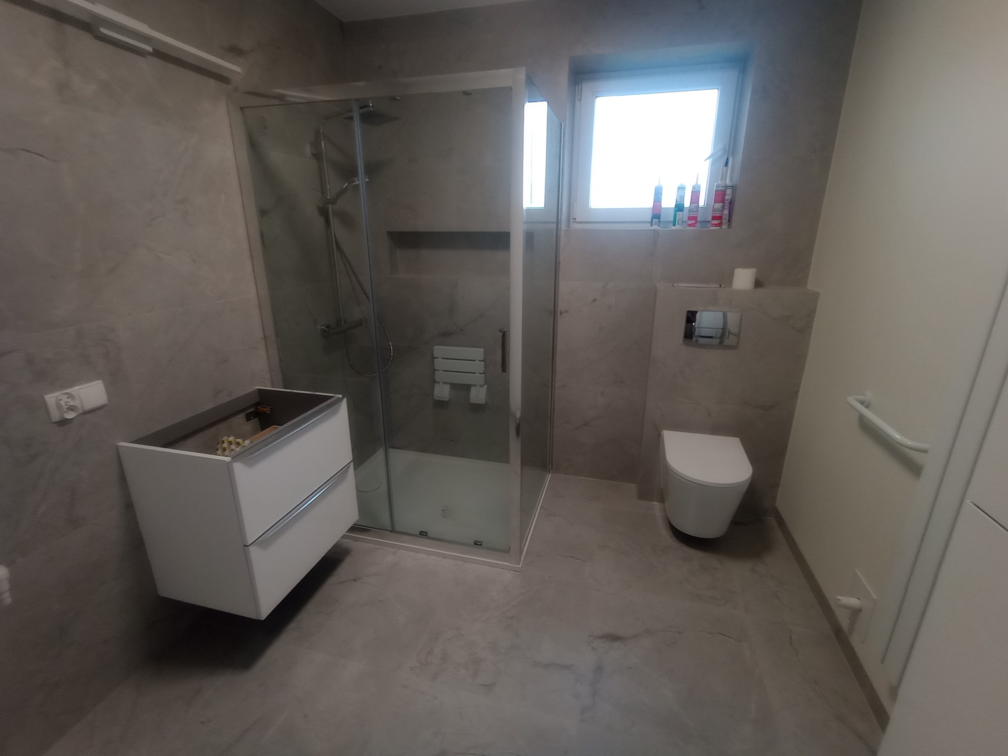 Wykończona łazienka w szarościach: kabina prysznicowa ze szklanymi drzwiami, podwieszana szafka z umywalką bez baterii, toaleta i okno z kosmetykami na parapecie.