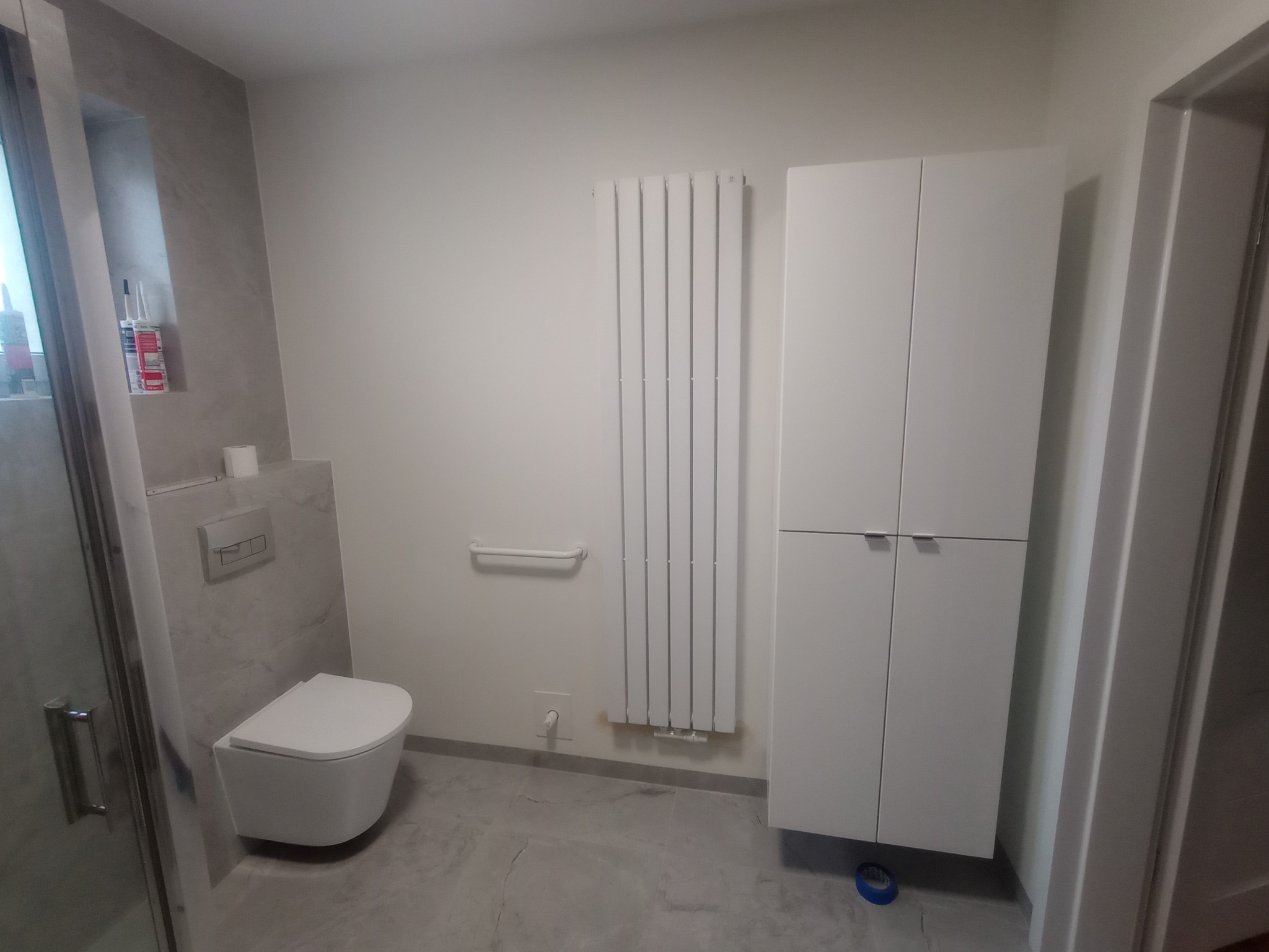 Nowoczesna łazienka po remoncie: podwieszana toaleta, grzejnik drabinkowy, biała szafka i szare płytki imitujące marmur. Minimalistyczny design, schludne wykończenie.