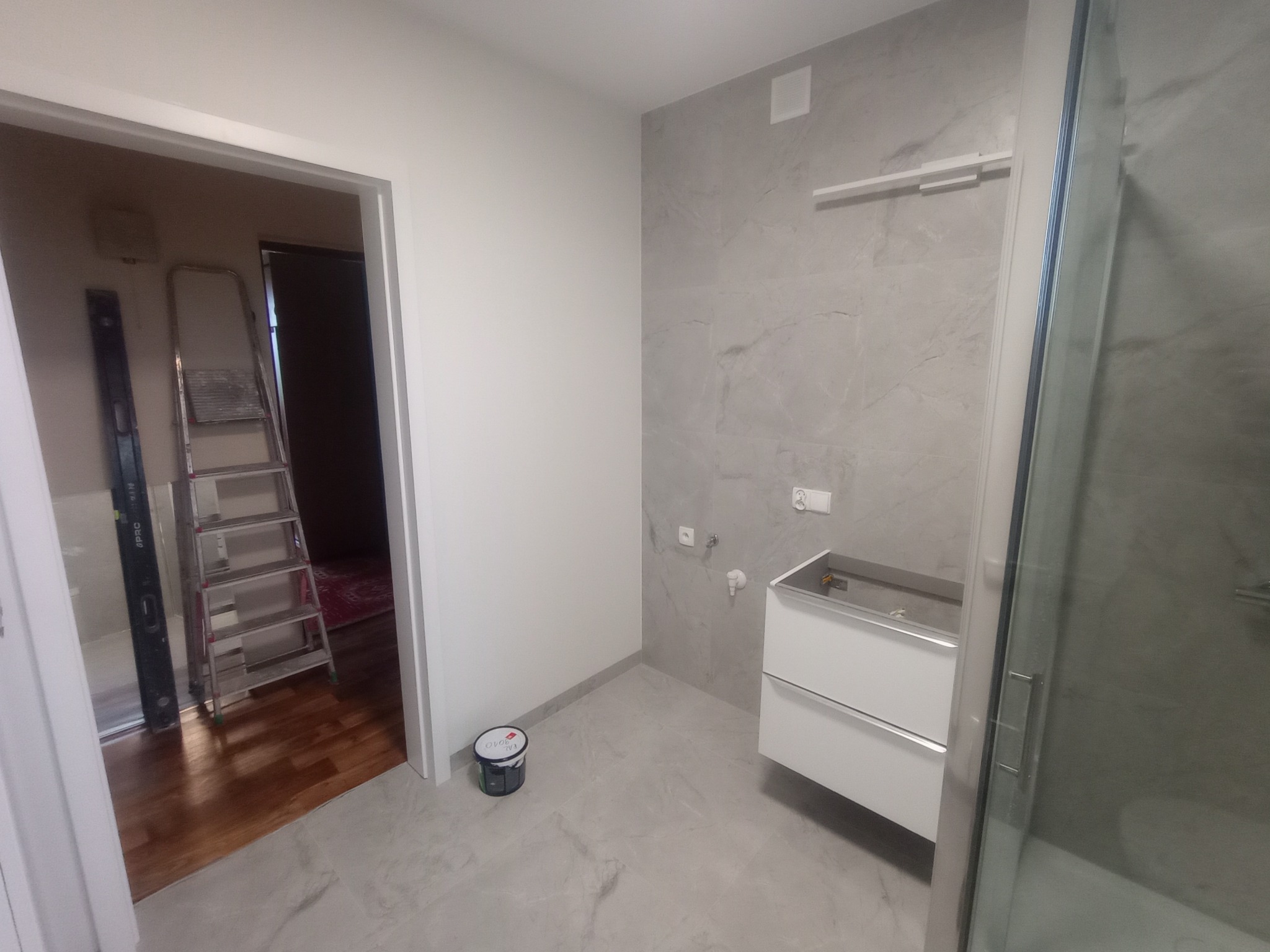 Wnętrze łazienki w trakcie prac wykończeniowych: widoczna umywalka, płytki imitujące marmur, drabina malarska w przejściu do innego pomieszczenia, wiadro z farbą.