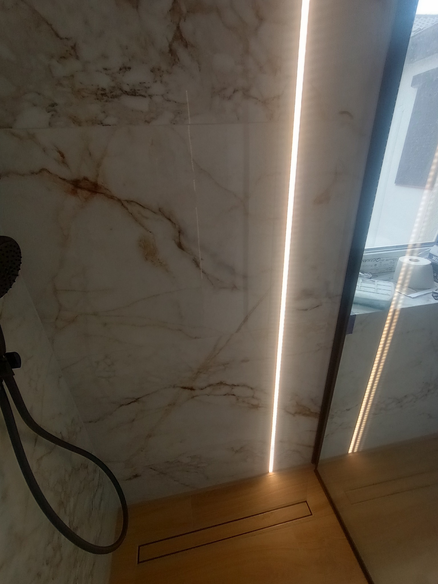 Wykończona łazienka: marmurowe płytki ścienne z oświetleniem LED, odpływ liniowy w drewnopodobnej posadzce i widoczny fragment słuchawki prysznicowej.