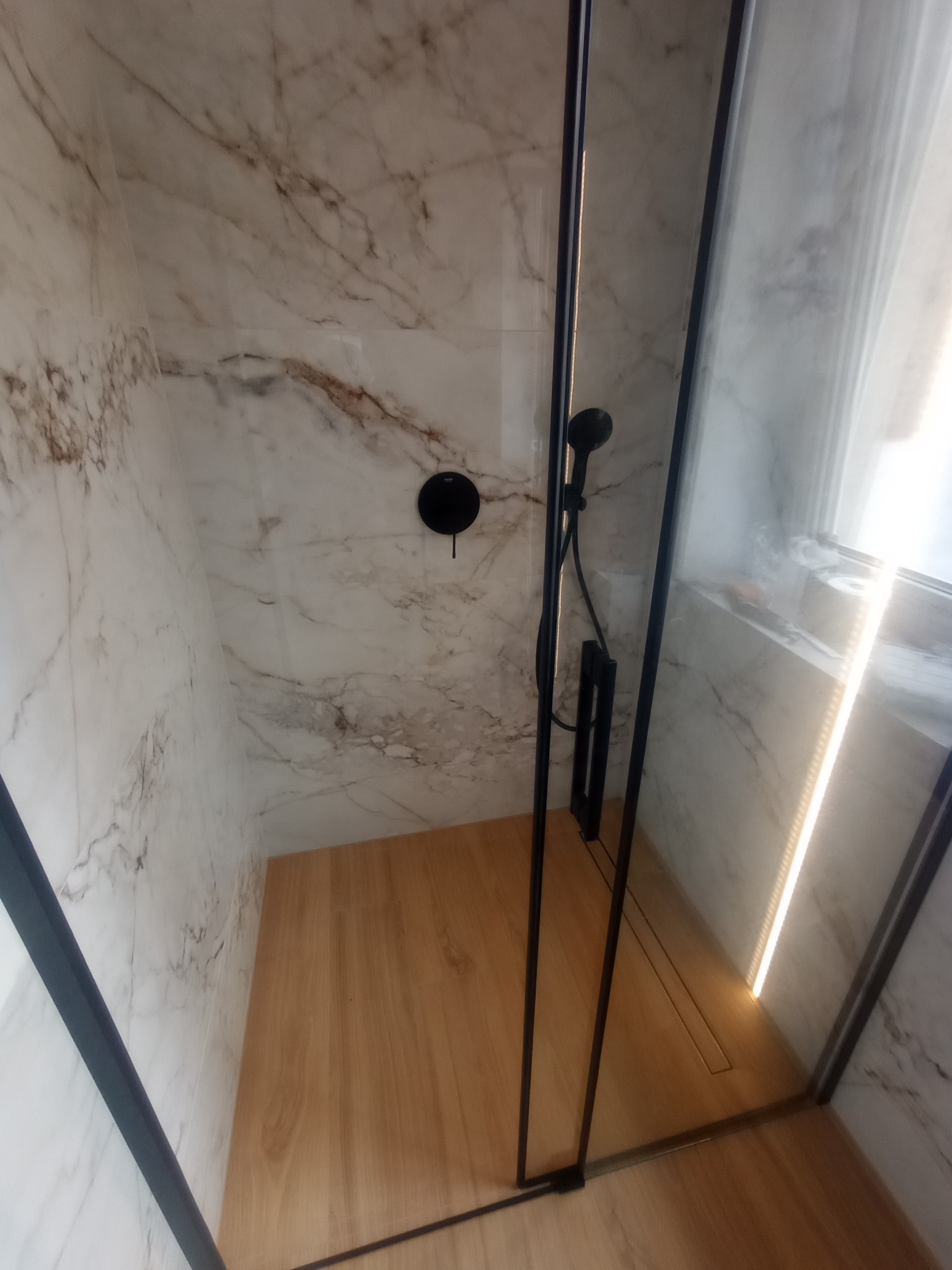 Nowoczesna kabina prysznicowa z czarnymi detalami, marmurowymi płytkami i drewnopodobną podłogą, oświetlona listwą LED. Minimalistyczny design.