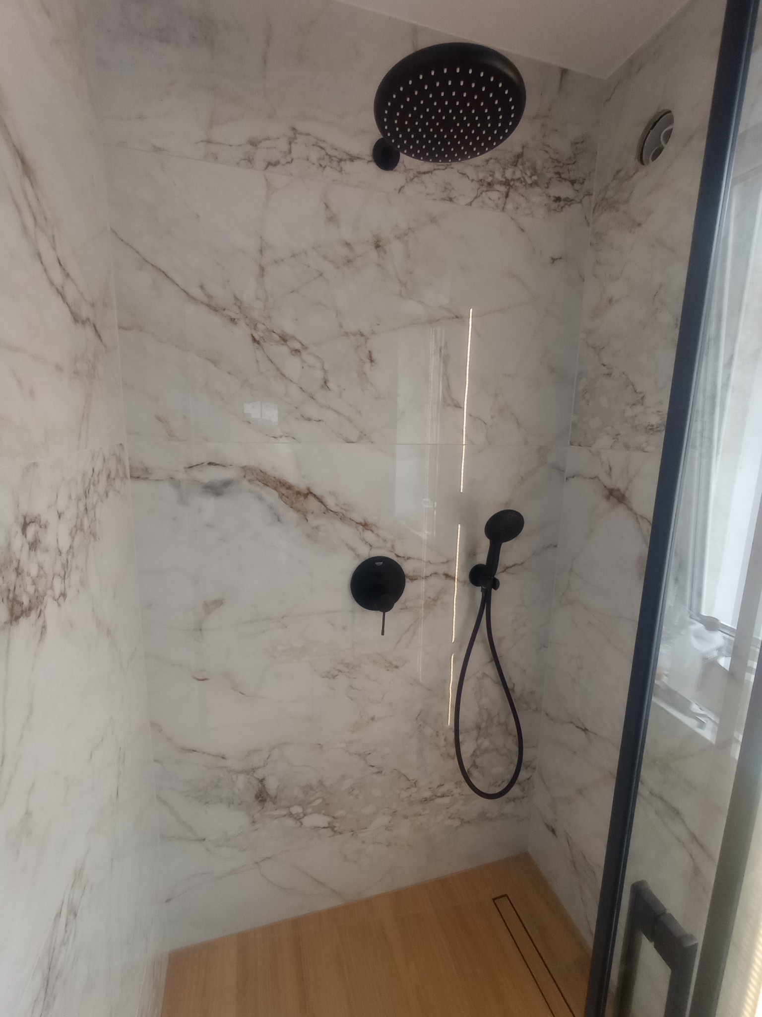 Nowoczesna kabina prysznicowa z marmurowymi płytkami, deszczownicą i drewnianym brodzikiem. Czarna armatura i minimalistyczny design.