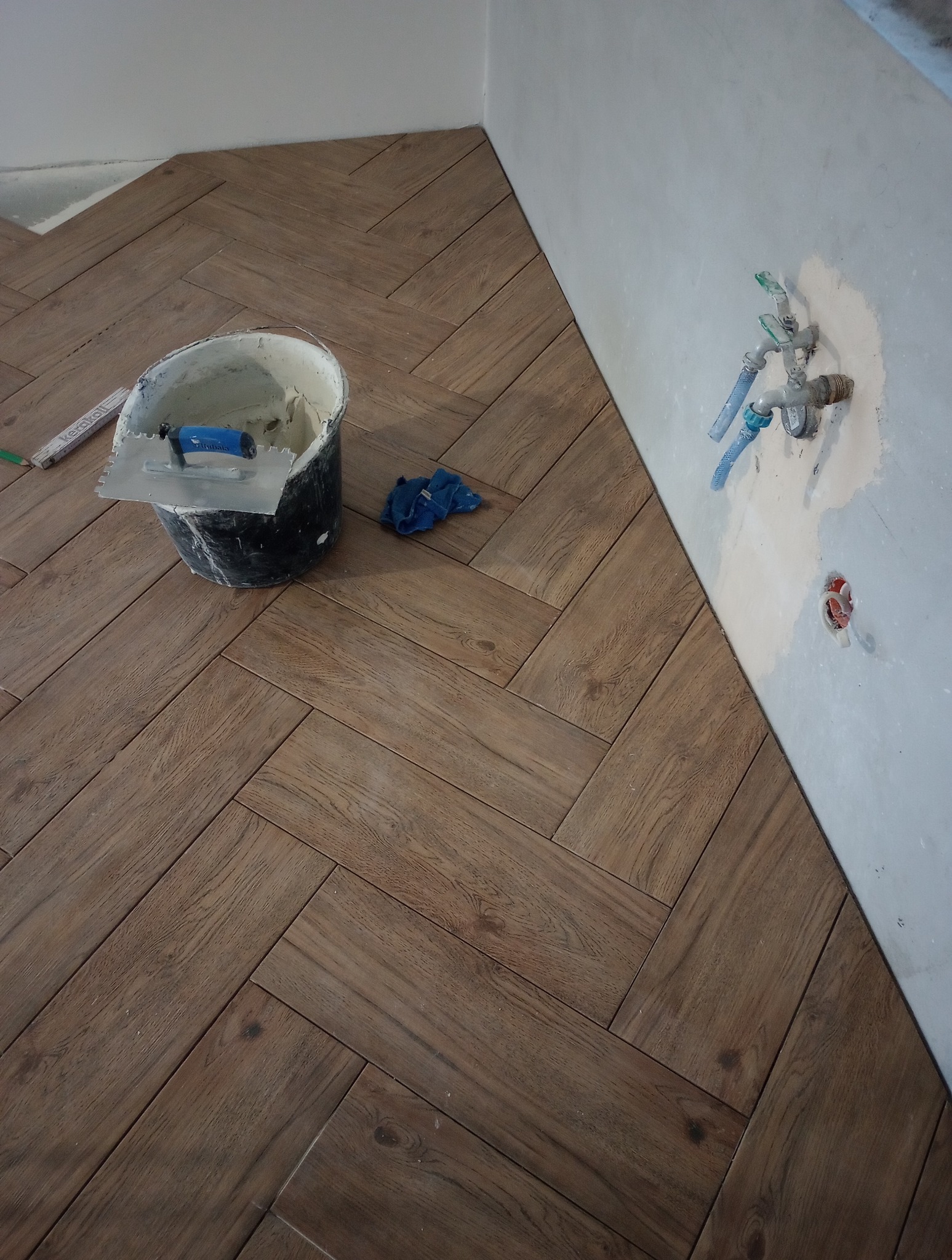 Ułożona podłoga z płytek imitujących drewno w jodełkę, wiadro z zaprawą i narzędzia. W tle fragment ściany z instalacją wodną. Remont łazienki w trakcie.