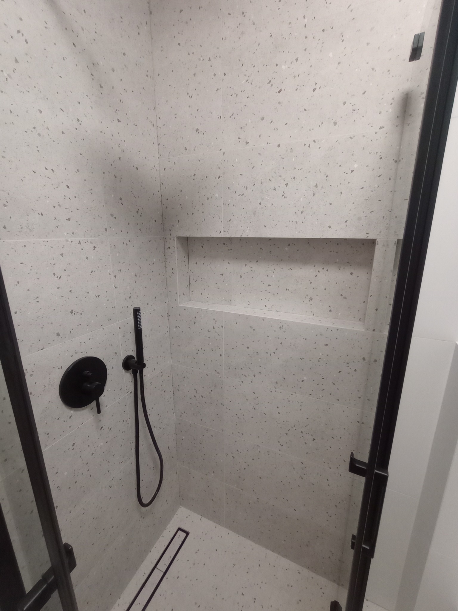 Nowoczesna kabina prysznicowa z czarną armaturą i odpływem liniowym, wykończona jasnymi płytkami w drobny wzór. W ścianie wnęka.