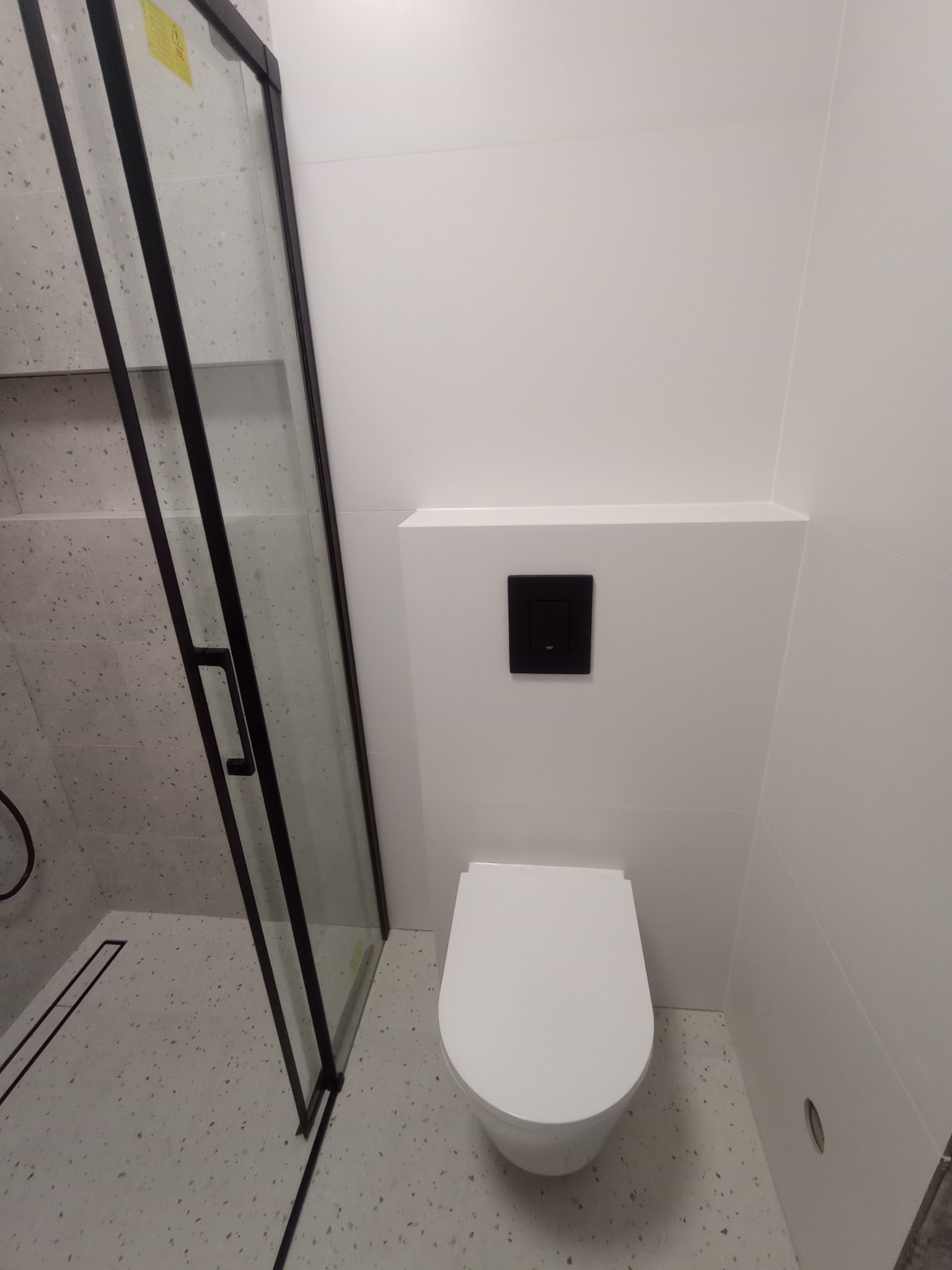 Nowoczesna łazienka w bieli z czarnymi akcentami: kabina prysznicowa typu walk-in i podwieszana toaleta. Minimalistyczny design i schludne wykończenie.