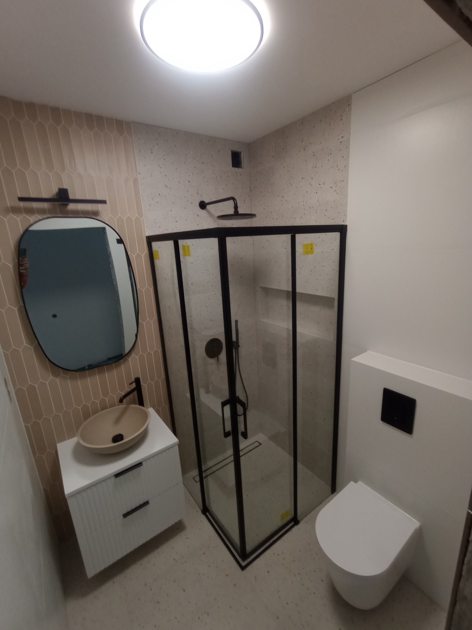 Nowoczesna łazienka z kabiną prysznicową typu walk-in z czarnymi ramami, owalnym lustrem i umywalką nablatową w beżowym odcieniu. Minimalistyczny design.