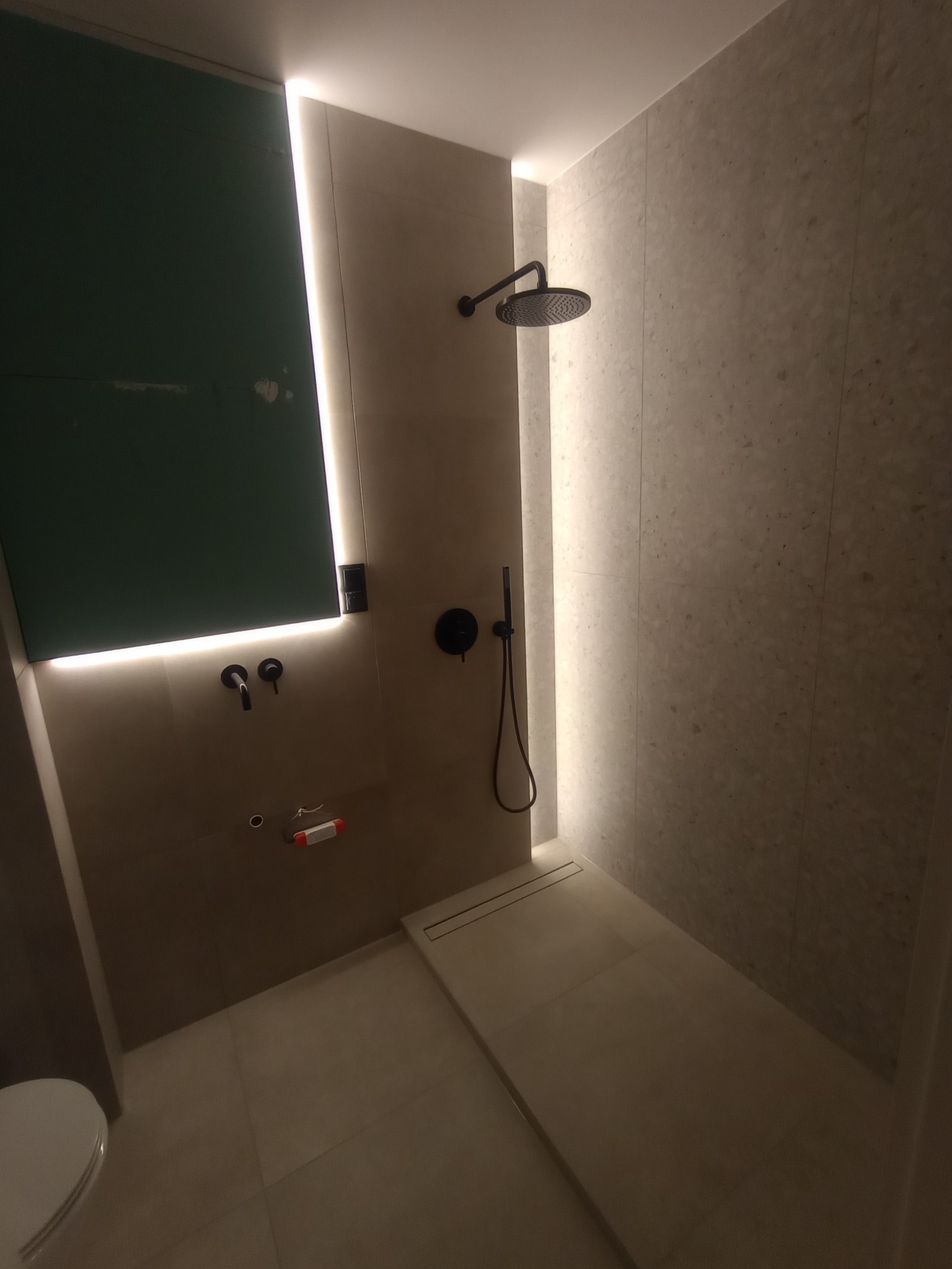 Nowoczesna łazienka w trakcie wykańczania: surowy gips, czarna armatura, jasne płytki i podświetlenie LED. Widoczny odpływ liniowy i fragment toalety.