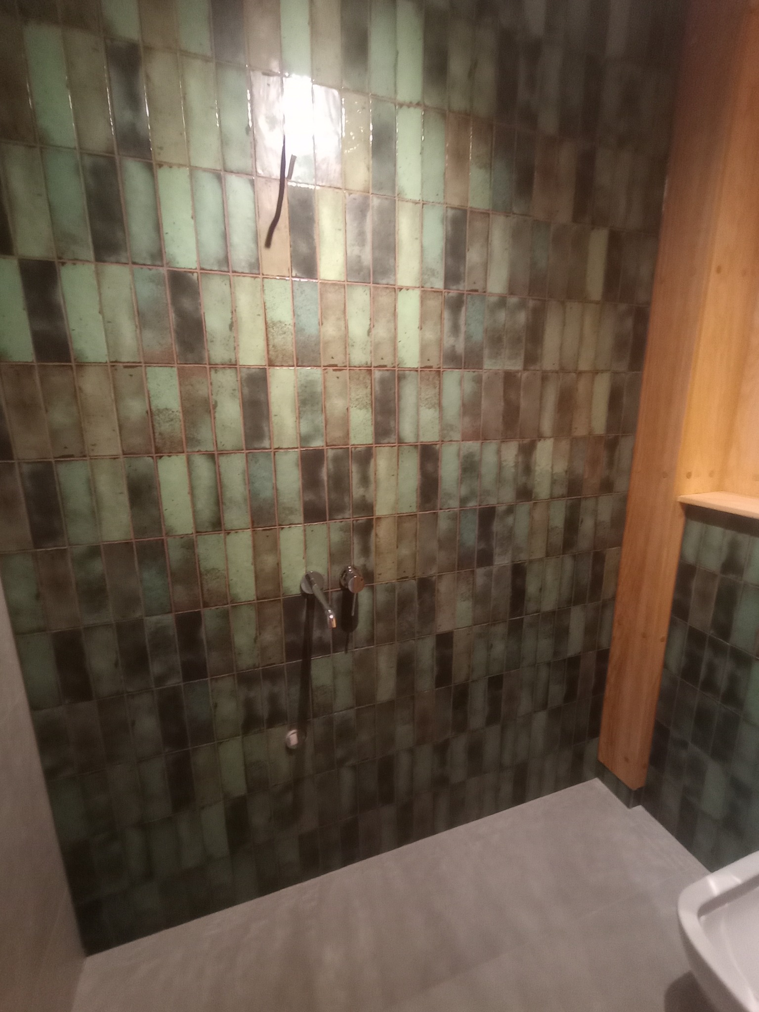 Wykończona łazienka z oryginalnymi, pionowo ułożonymi, zielono-brązowymi płytkami. Widoczna armatura i drewniany element wykończeniowy.