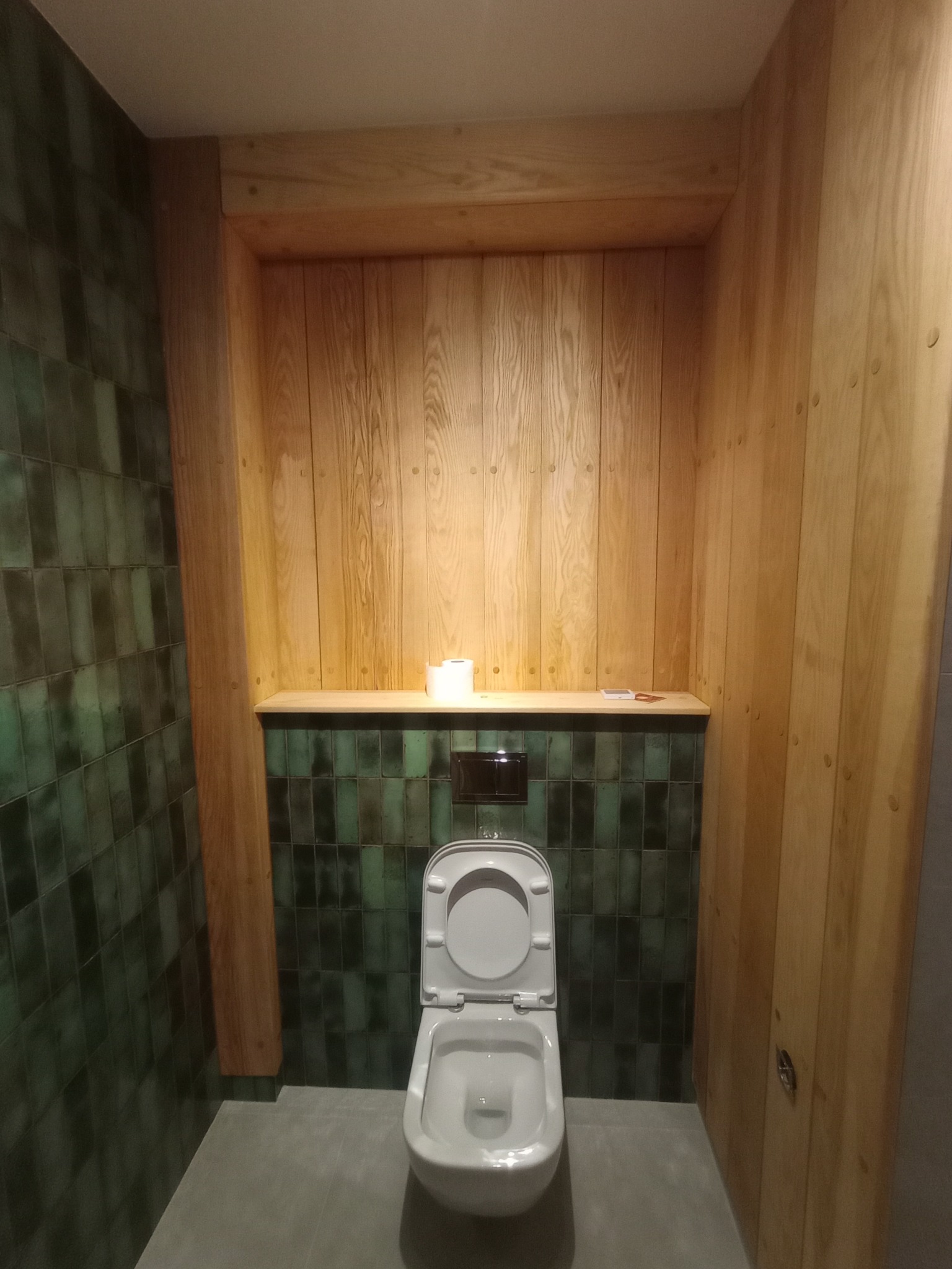 Nowoczesna toaleta z drewnianą zabudową i zielonymi płytkami. Minimalistyczny design, ciepłe oświetlenie. Ukończona realizacja wykończenia łazienki.