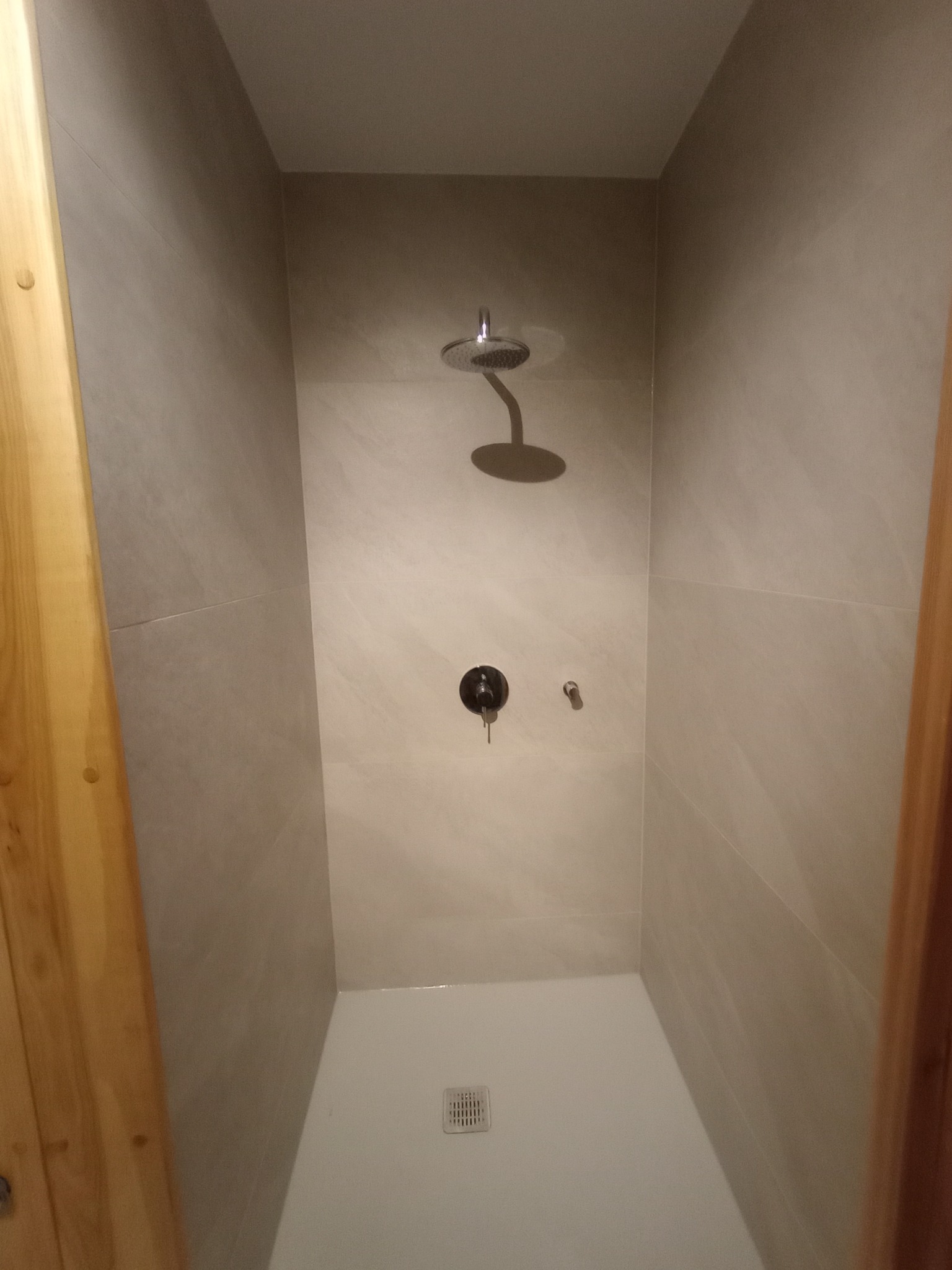 Nowoczesna kabina prysznicowa walk-in z deszczownicą i ciemną armaturą na tle beżowych płytek. Widoczny drewniany element wykończenia.