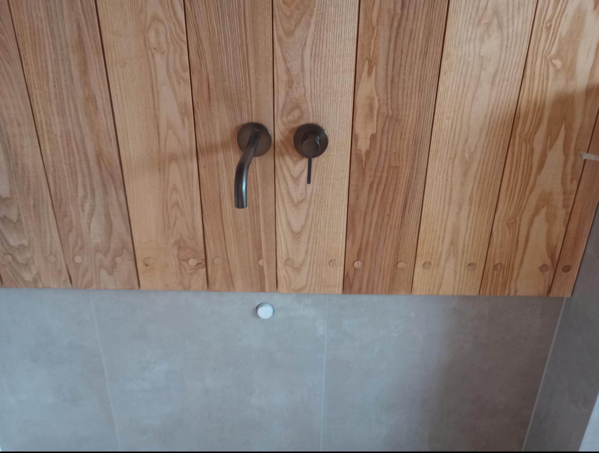 Drewniana ściana z minimalistyczną, czarną armaturą w łazience. Widoczne naturalne usłojenie drewna i subtelne detale wykończenia. Nowoczesny design.