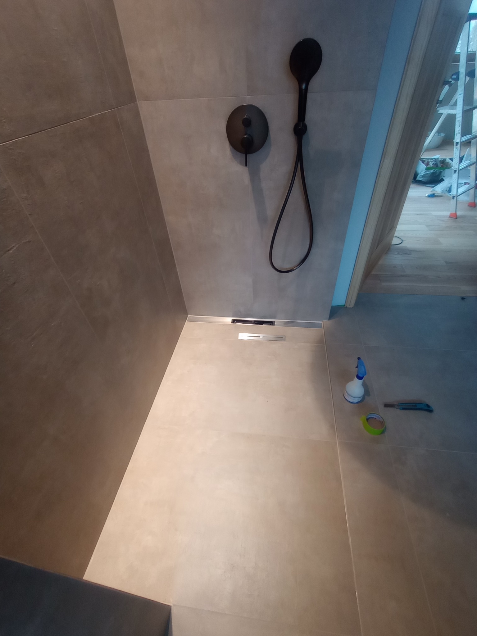 Nowoczesna kabina prysznicowa z odpływem liniowym, wykończona szarą płytką. Widoczne narzędzia i akcesoria montażowe. Minimalistyczny design.