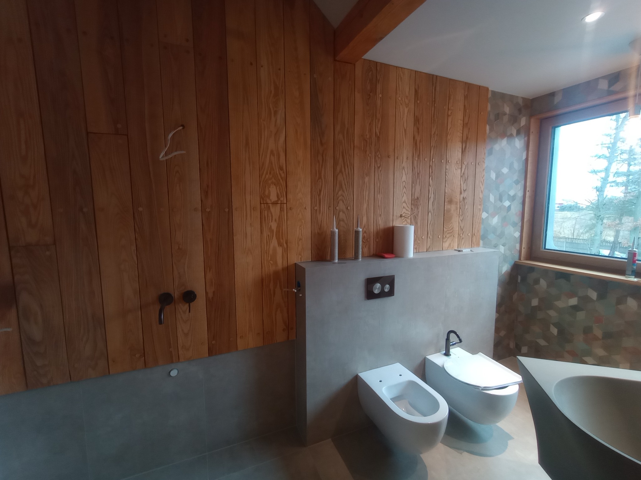 Nowoczesna łazienka z drewnianą okładziną ściany, minimalistyczną armaturą i geometrycznym wzorem na ścianie przy oknie. Widok na umywalkę, bidet i toaletę.