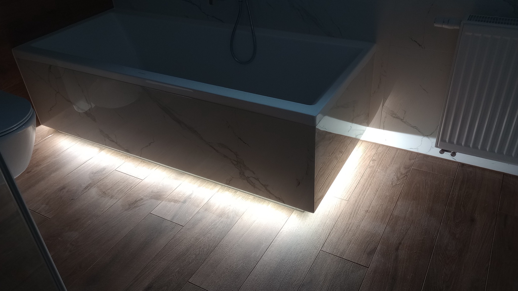 Nowoczesna łazienka z podświetloną wanną w marmurze i drewnopodobną podłogą. Grzejnik na ścianie z marmurowym wzorem. Częściowo widoczna toaleta.