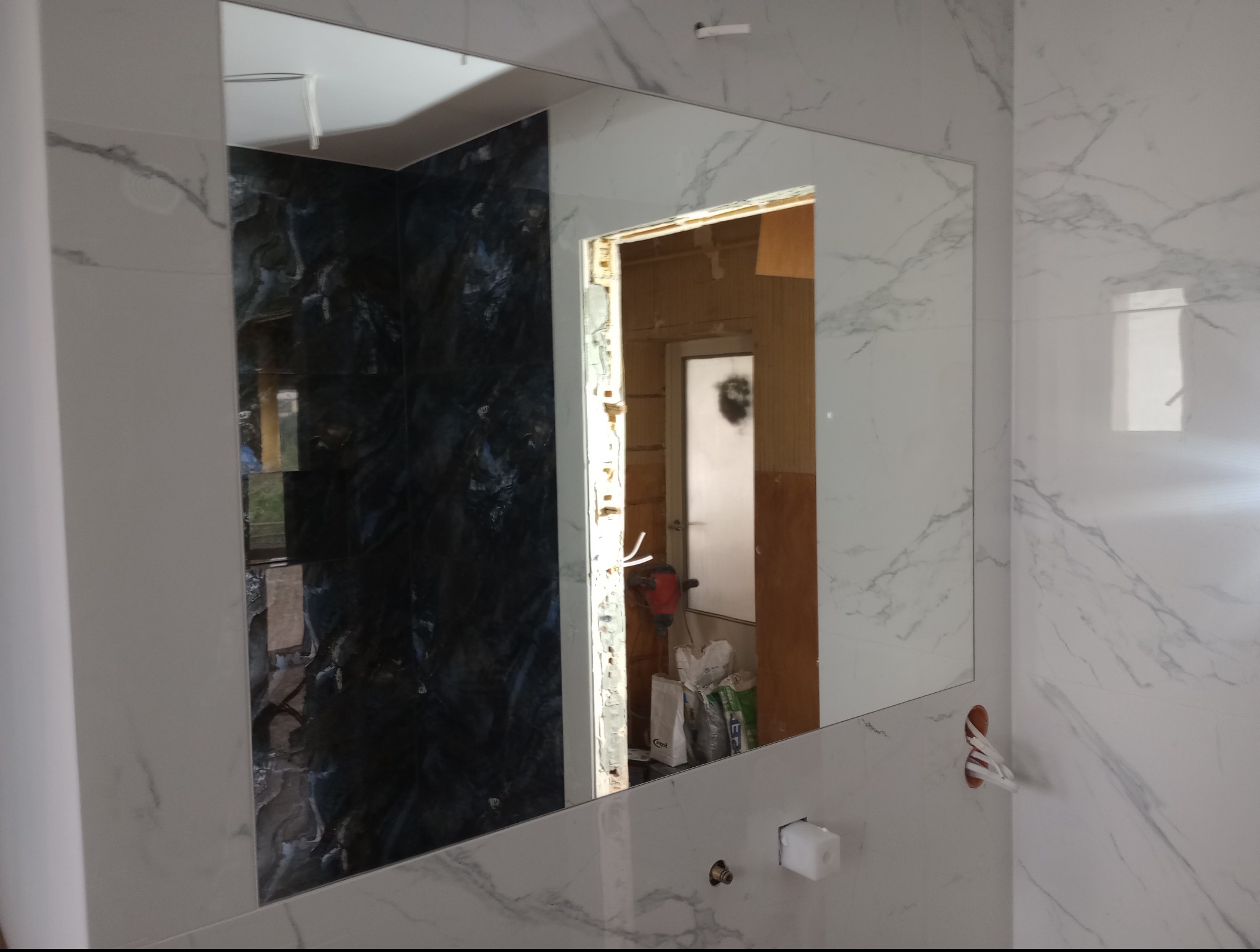 Łazienka w trakcie prac wykończeniowych: białe płytki imitujące marmur, lustro bez ramy, widoczne instalacje i narzędzia, fragment sąsiedniego pomieszczenia w odbiciu.