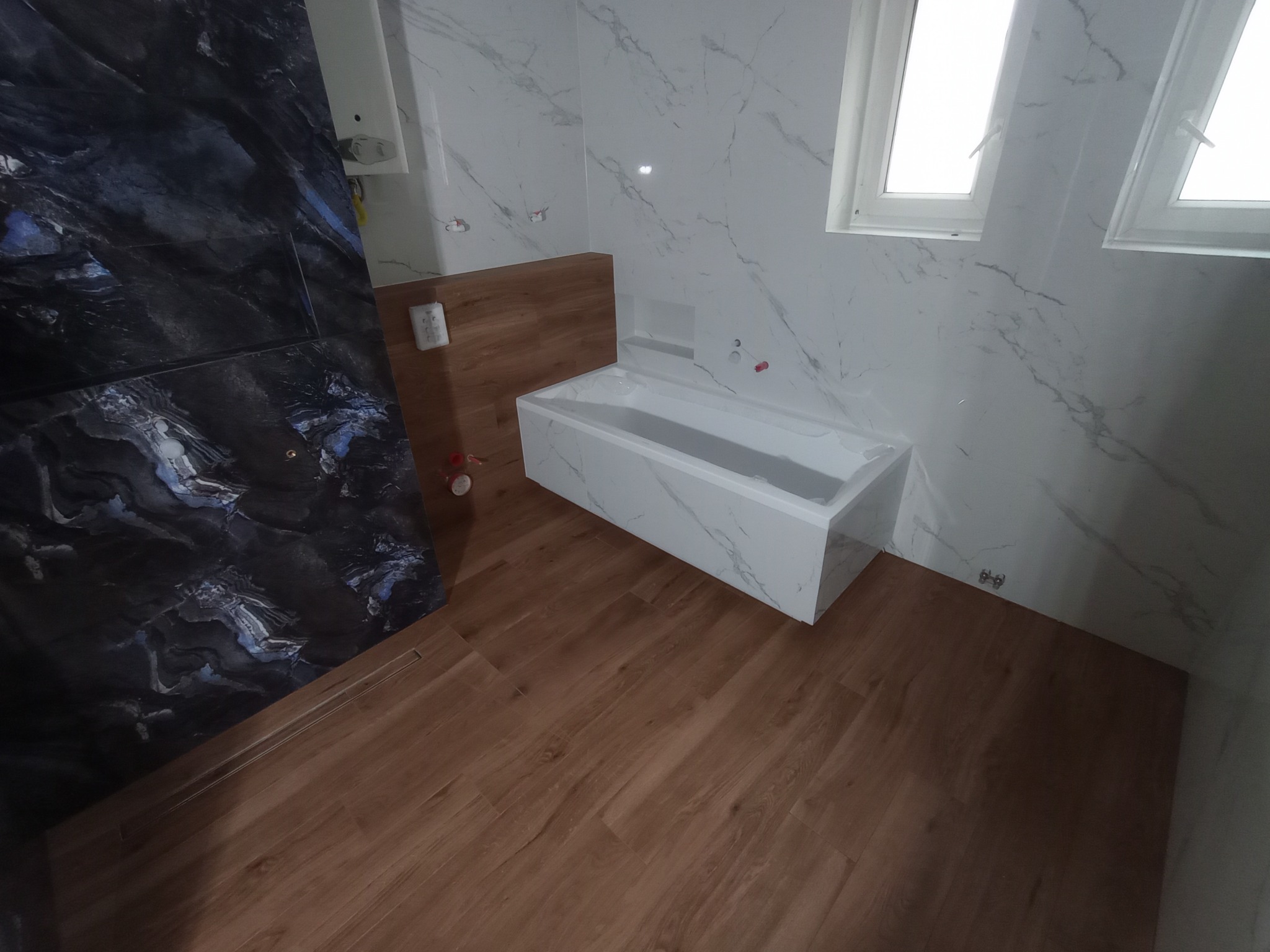 Nowoczesna łazienka z wanną w zabudowie, marmurowe płytki ścienne i podłoga imitująca drewno. Uwagę przykuwa kontrast ciemnej ściany z jasnymi elementami.