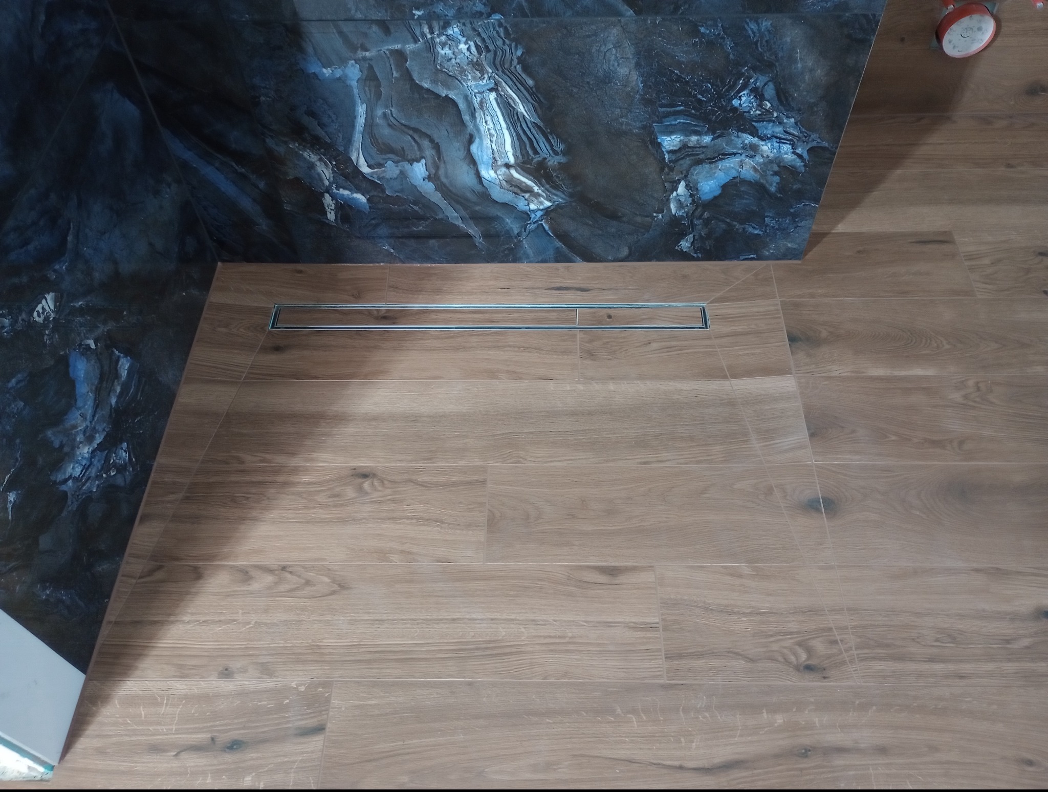 Nowoczesna łazienka z drewnopodobną podłogą i odpływem liniowym, oraz ścianą z płytek imitujących marmur w odcieniach granatu i szarości.