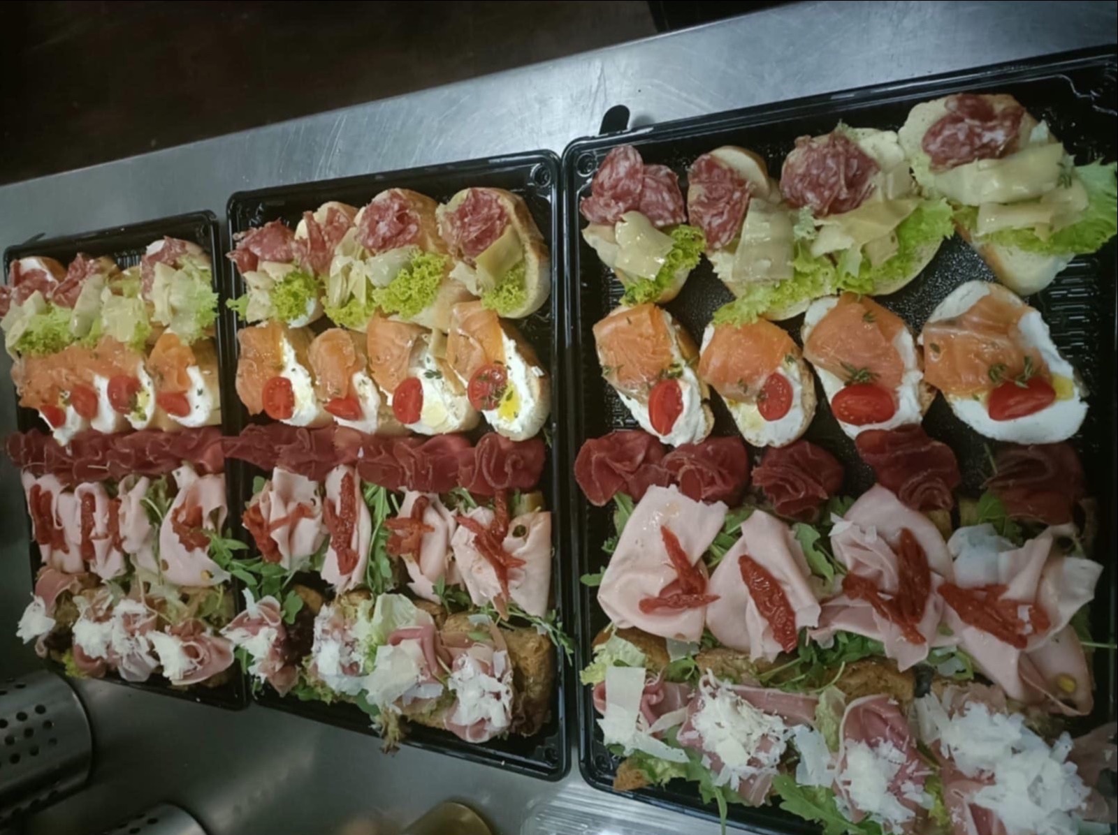 Dwie czarne tace z różnorodnymi kanapkami: z łososiem, szynką parmeńską, salami i serem, udekorowane świeżymi warzywami. Widok z góry.