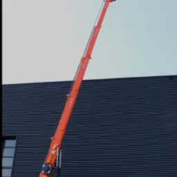 Pomarańczowy dźwig gąsienicowy, wysięgnik w górze, stabilizatory rozłożone, na tle ciemnej hali. Widoczne detale konstrukcji i mechanizmów.