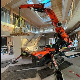 WEŹTOPODNIEŚ.PL Łukasz Dąbrowski - Pomarańczowy mini dźwig gąsienicowy Jansen S5000 podnosi paletę z materiałami wewnątrz budynku z drewnianą konstrukcją i schodami.